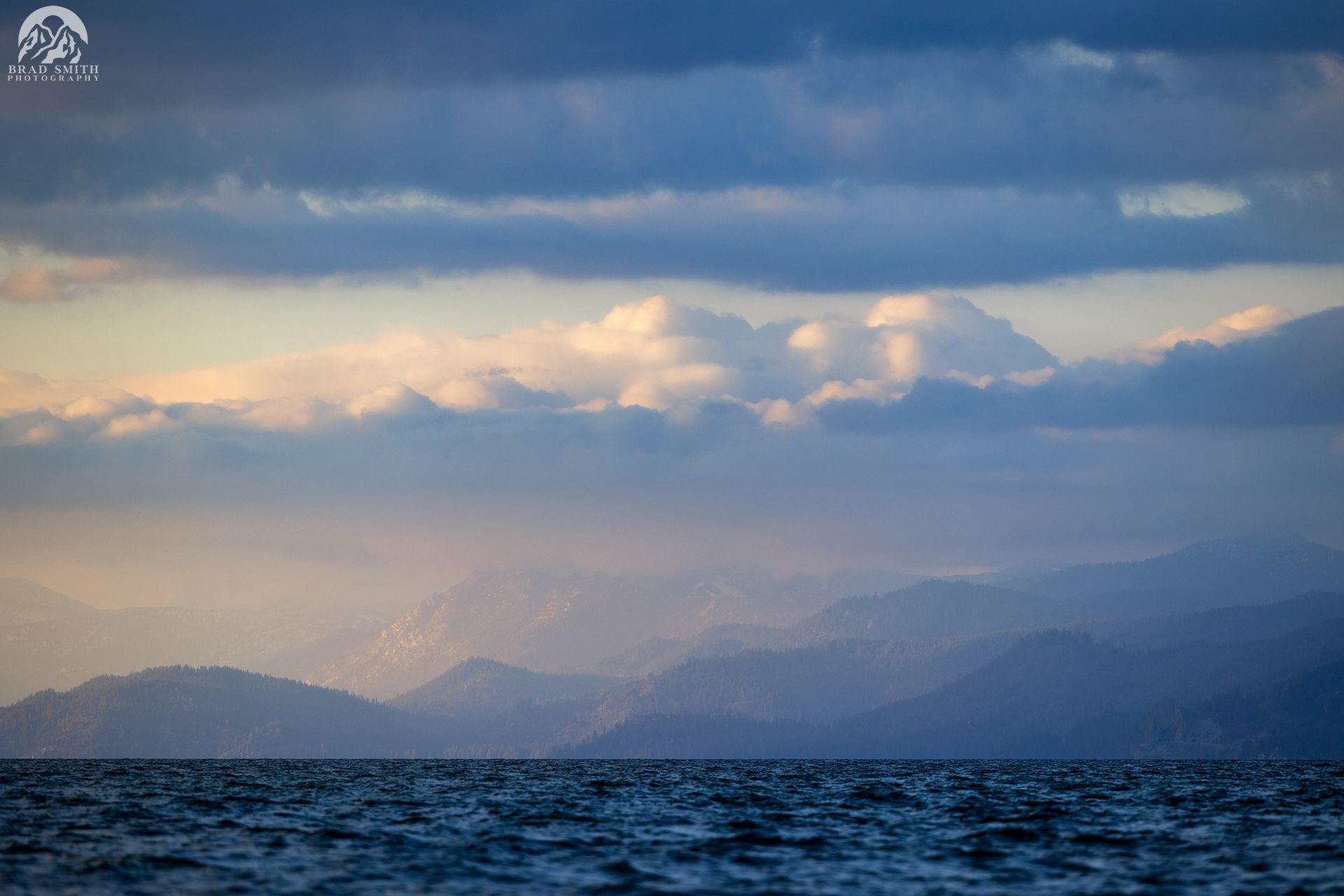 Stormy ocean under cloudy sky with layered mountains in the distance and soft sunset light
