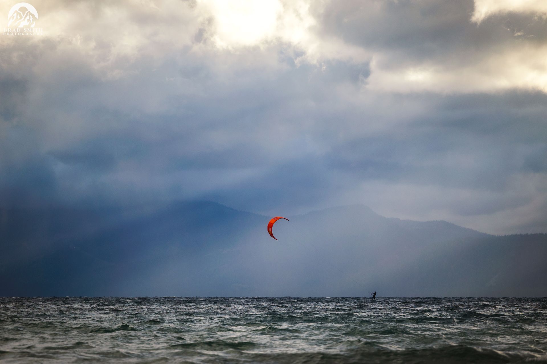Red kite surfacing above choppy water under dramatic cloudy sky near distant mountains