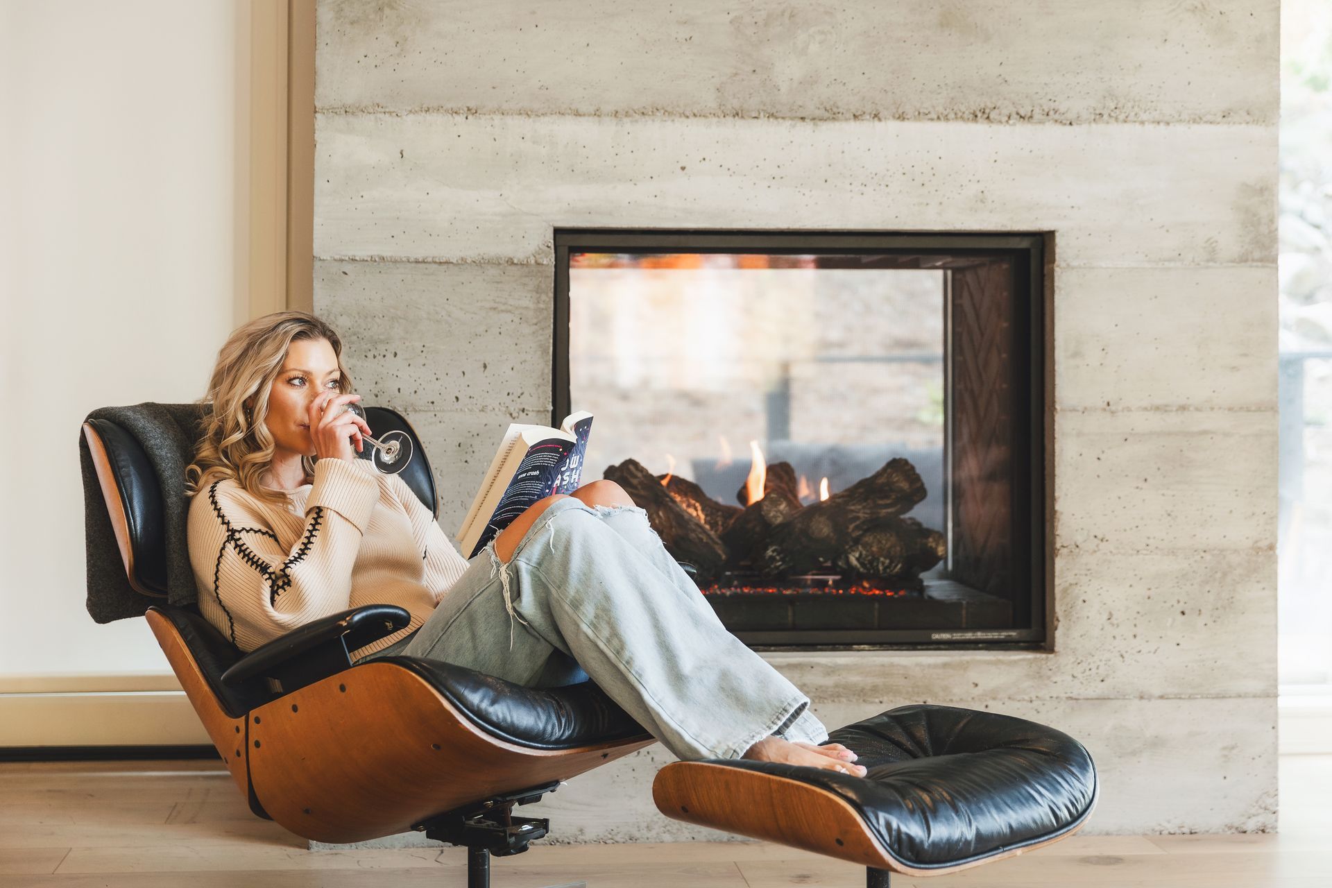Person relaxing in a black lounge chair beside a modern fireplace, reading a magazine.