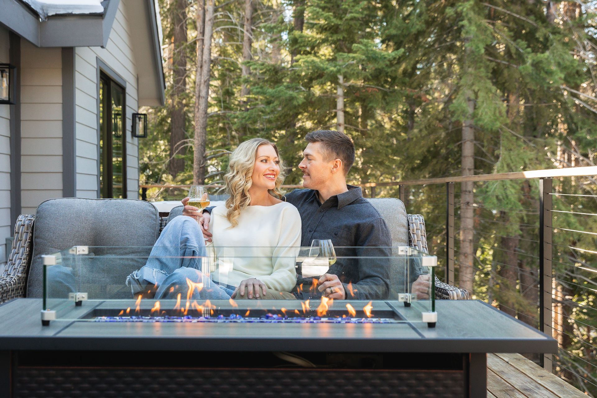Couple relaxing by an outdoor fire pit on a deck surrounded by trees and a house