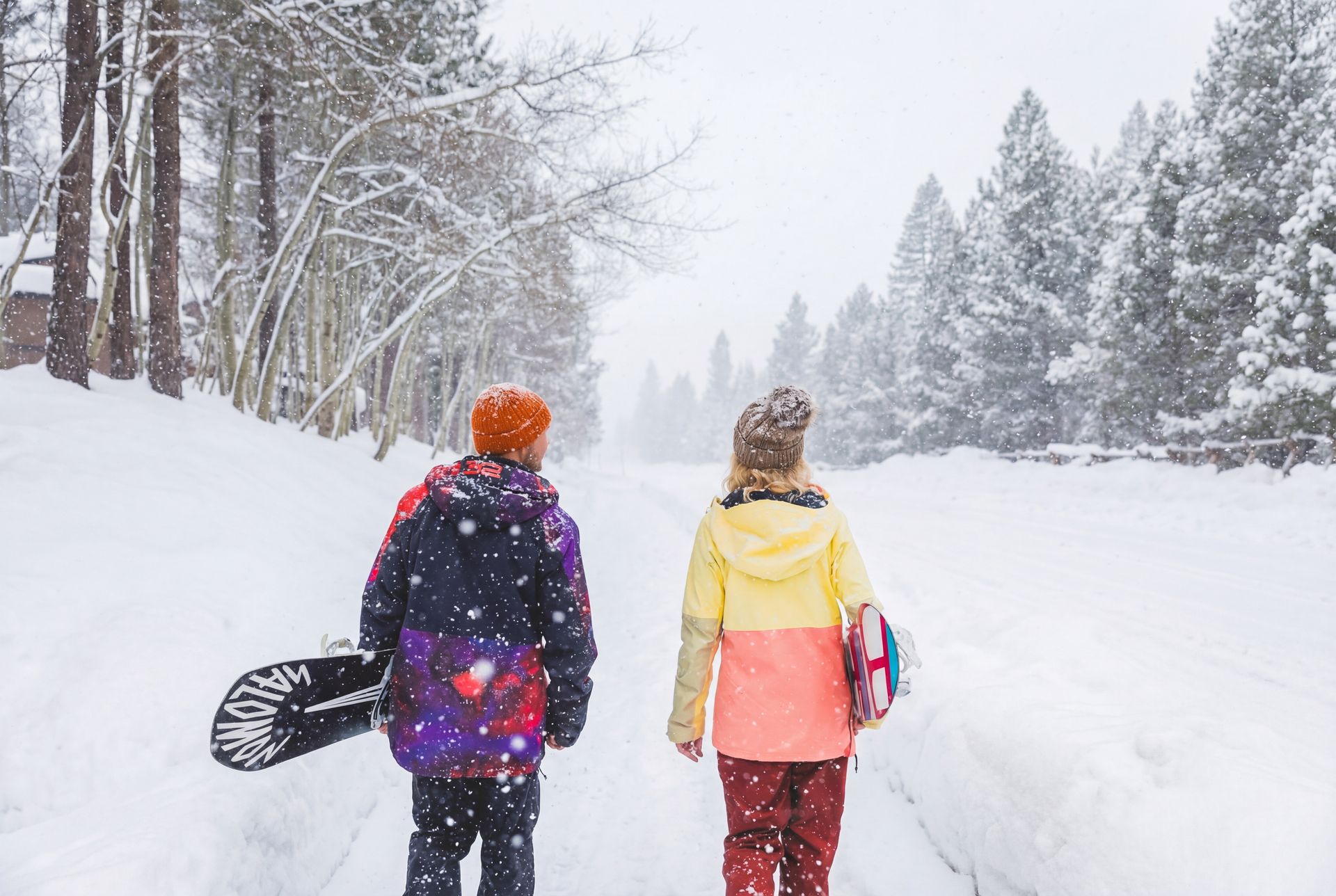Two people walking through a snowy forest, carrying a snowboard on a winter trail