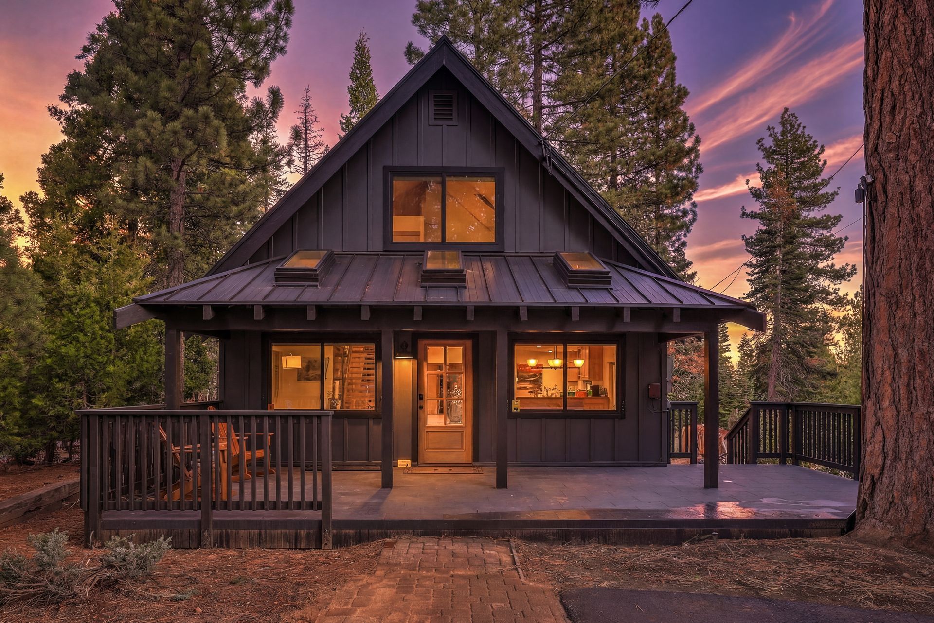 Cozy cabin at dusk with warm lit windows, surrounded by tall pine trees under a pink-purple sky