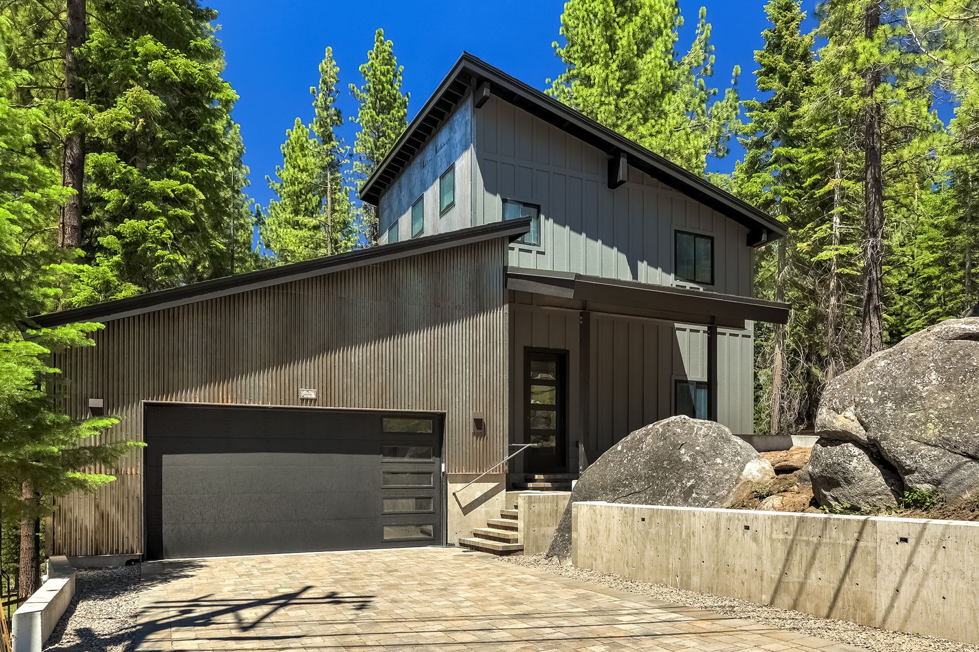 Modern gray house with garage beside large rocks and pine trees under a blue sky