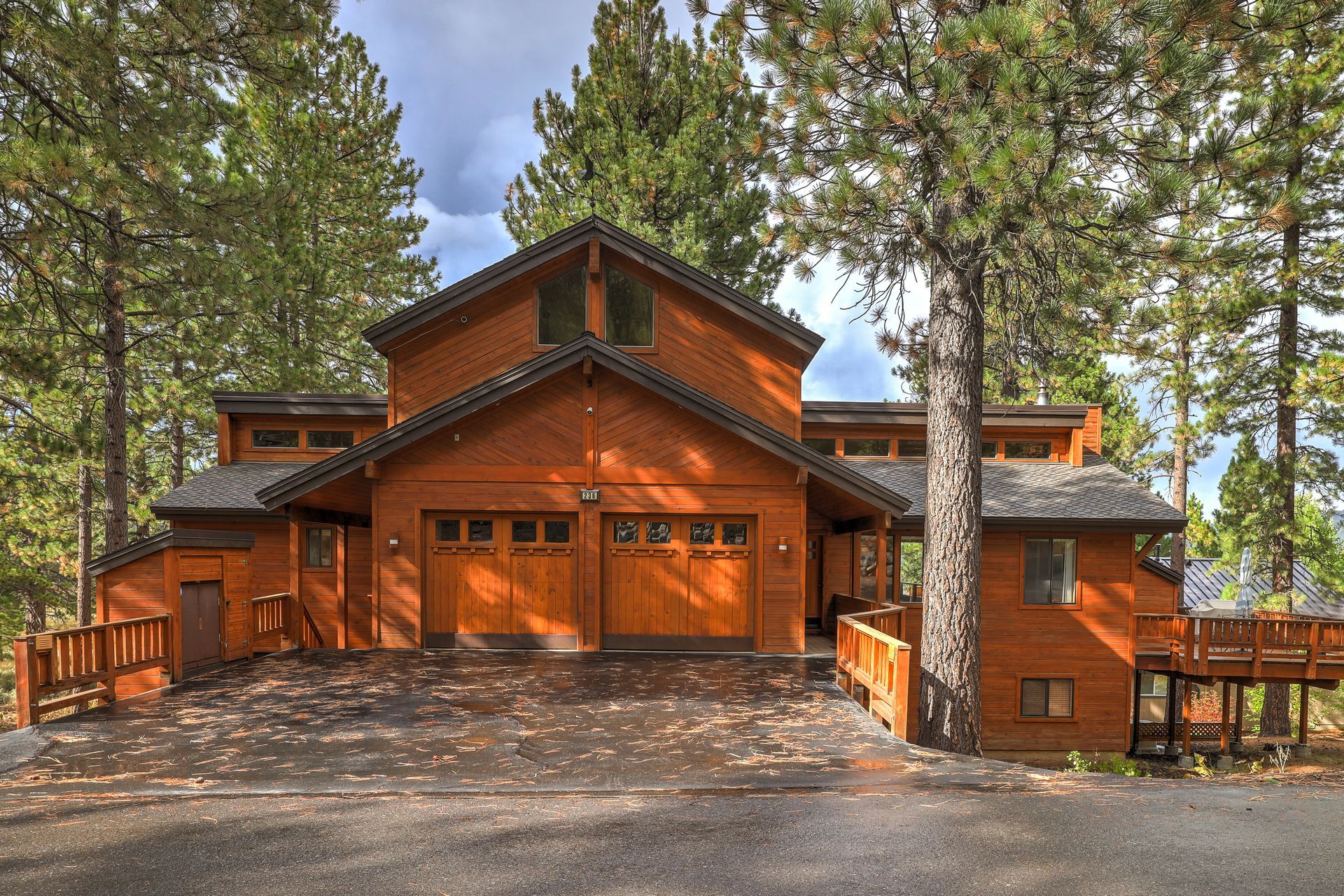 Rustic wooden cabin with a steep roof, framed by tall pine trees and a gravel driveway.