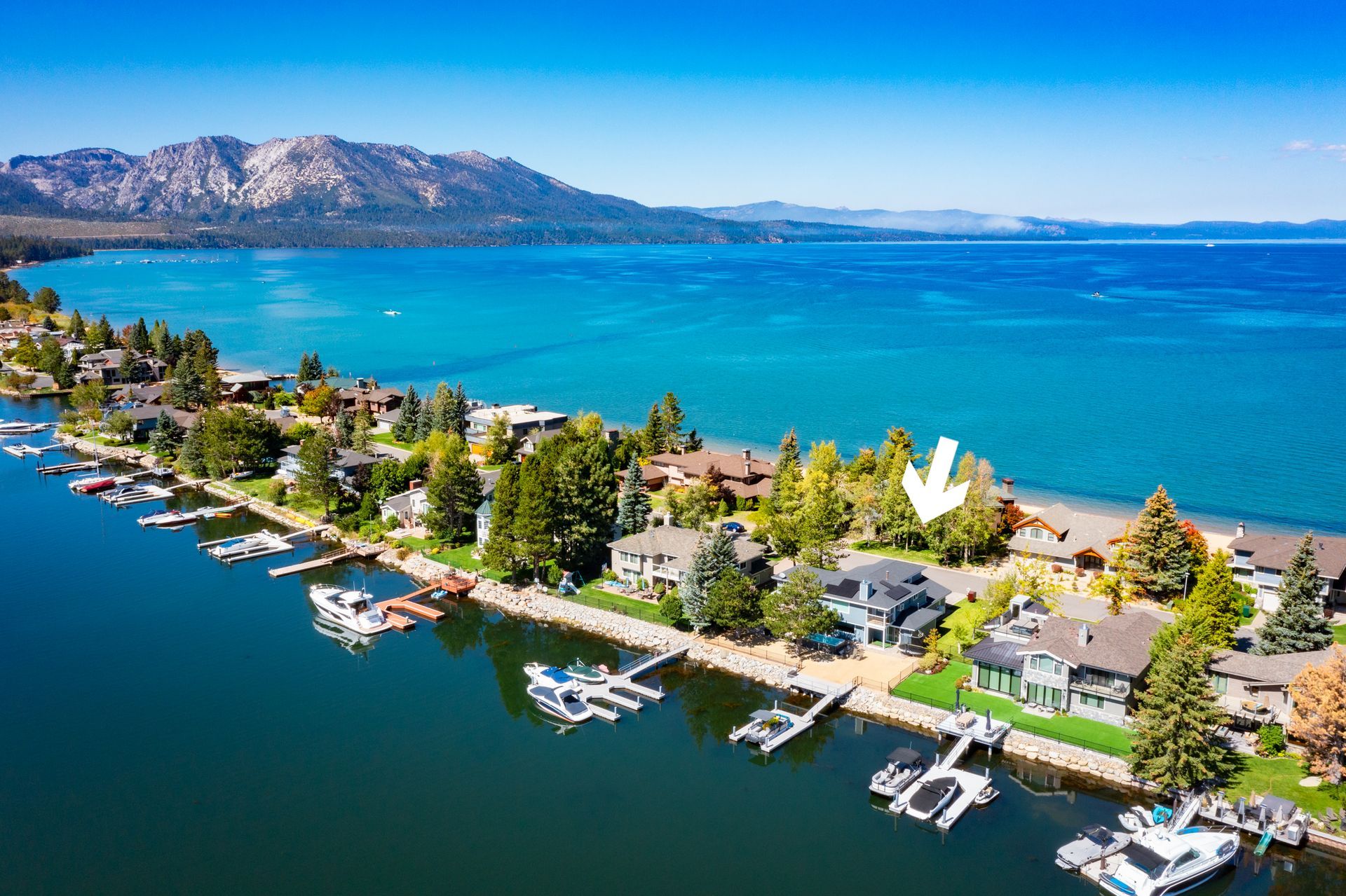 Aerial view of a lakeside neighborhood with docks, boats, and a clear blue lake backed by mountains.