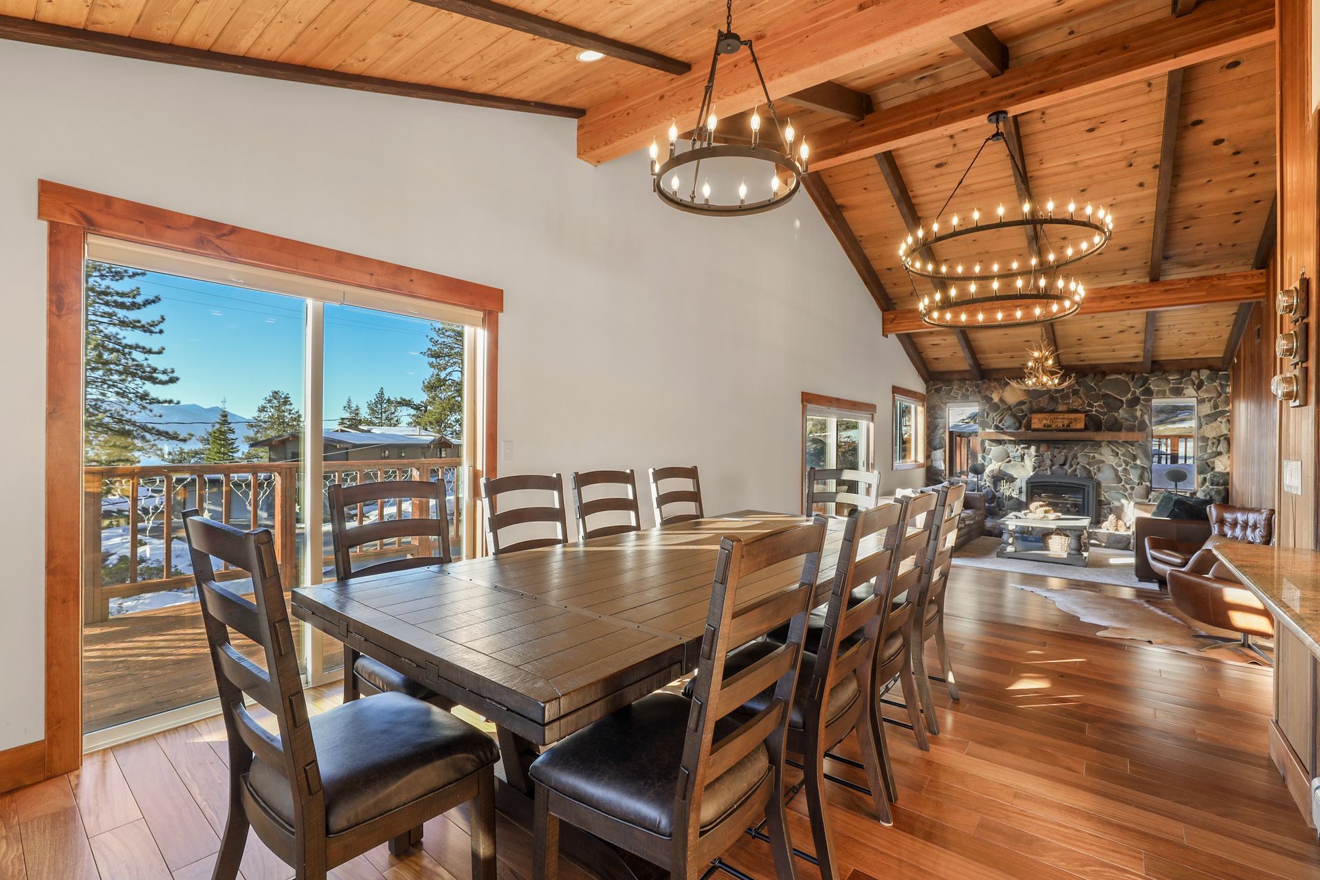 Rustic dining room with wood beams, long table, chandeliers, and large windows overlooking a deck and trees