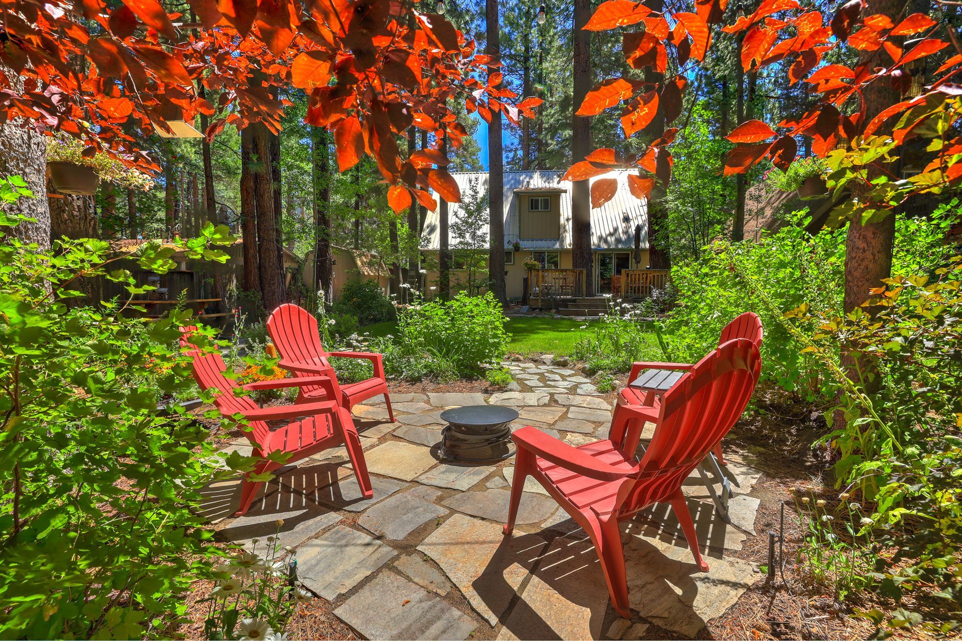 Red Adirondack chairs around a stone fire pit in a leafy backyard, framed by bright orange autumn leaves