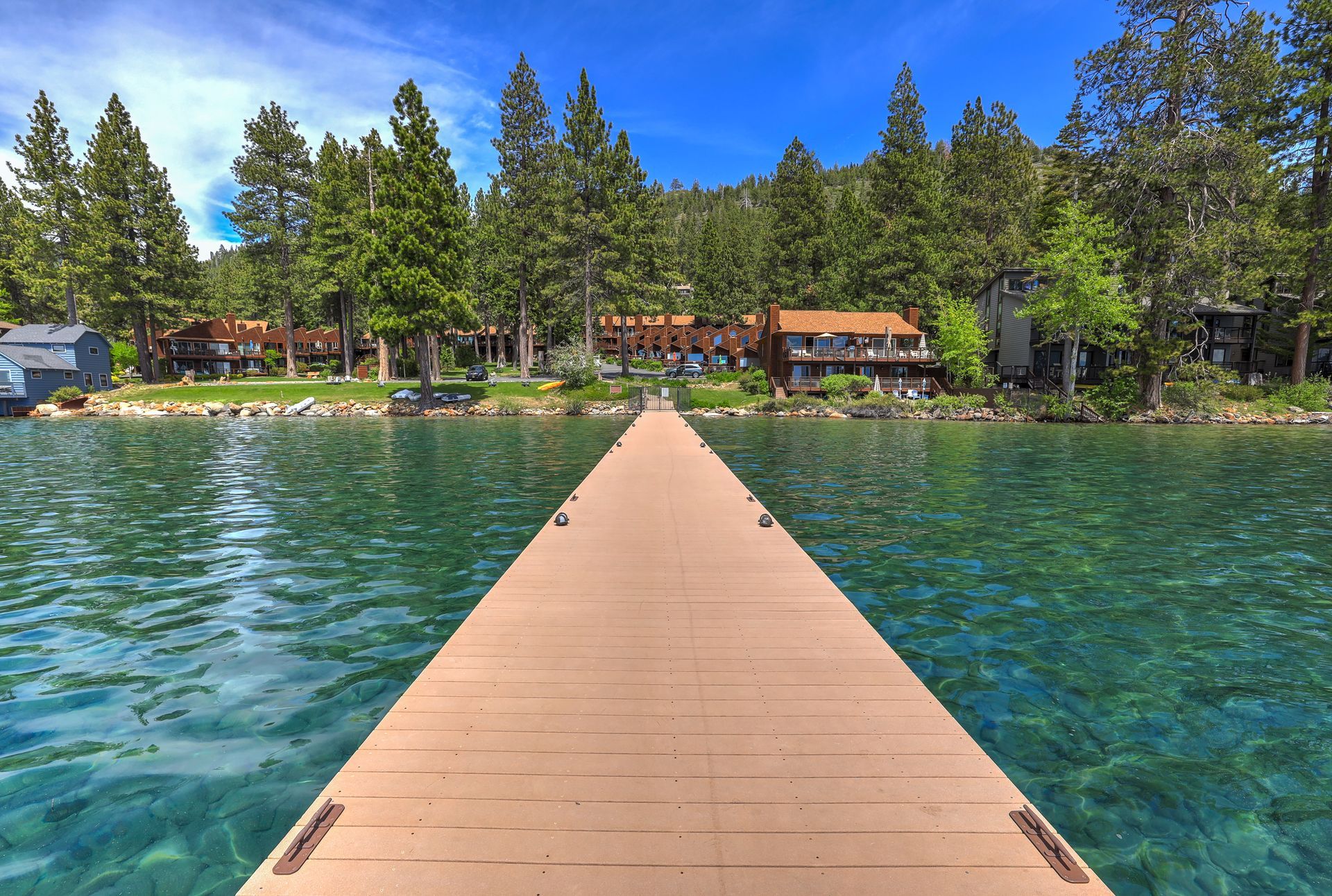 Wooden dock leading to a lakeside resort with turquoise water, pine trees, and cabins under a blue sky