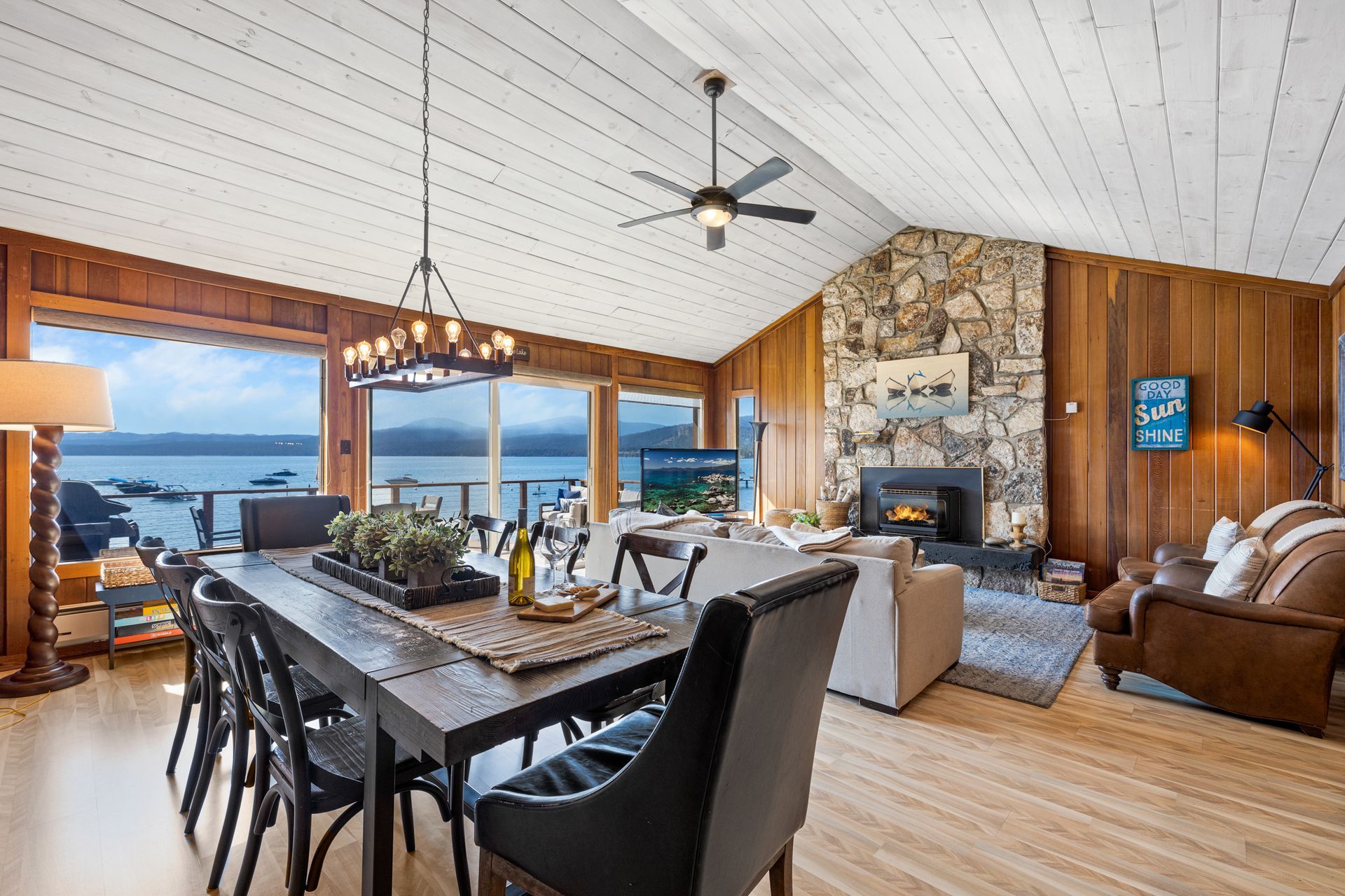 Bright open living room and dining area with lake views, wood paneling, stone fireplace, and leather chairs