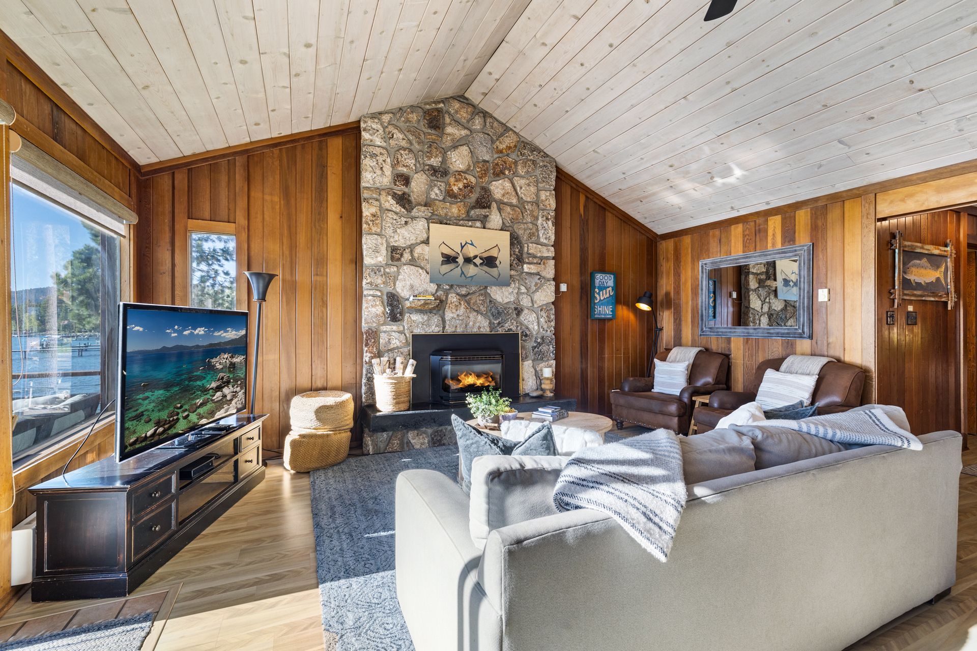 Cozy rustic living room with stone fireplace, wood-paneled walls, and gray sectional sofa.