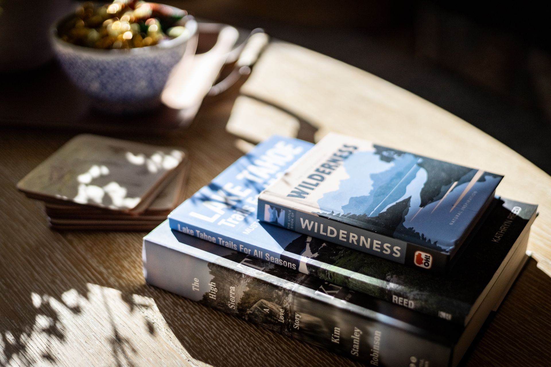 Stacked books on a sunlit wooden table beside a bowl of fruit and a folded napkin