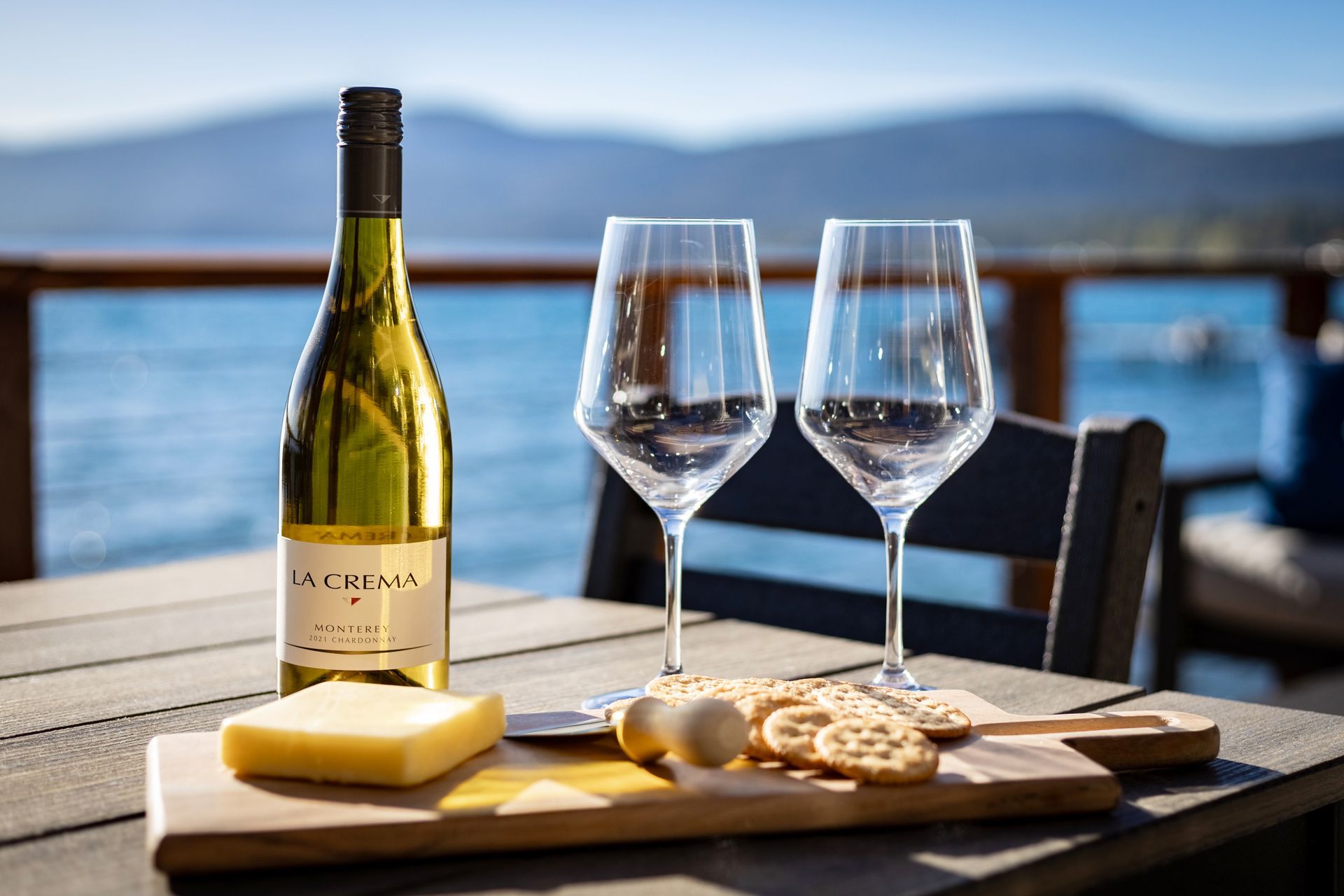 Wine bottle, two empty glasses, cheese and crackers on a tray by a lakeside table.