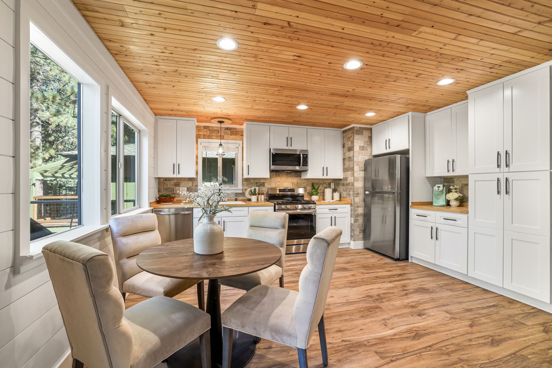 Bright kitchen with white cabinets, wood ceiling, stainless appliances, and a dining table by large windows
