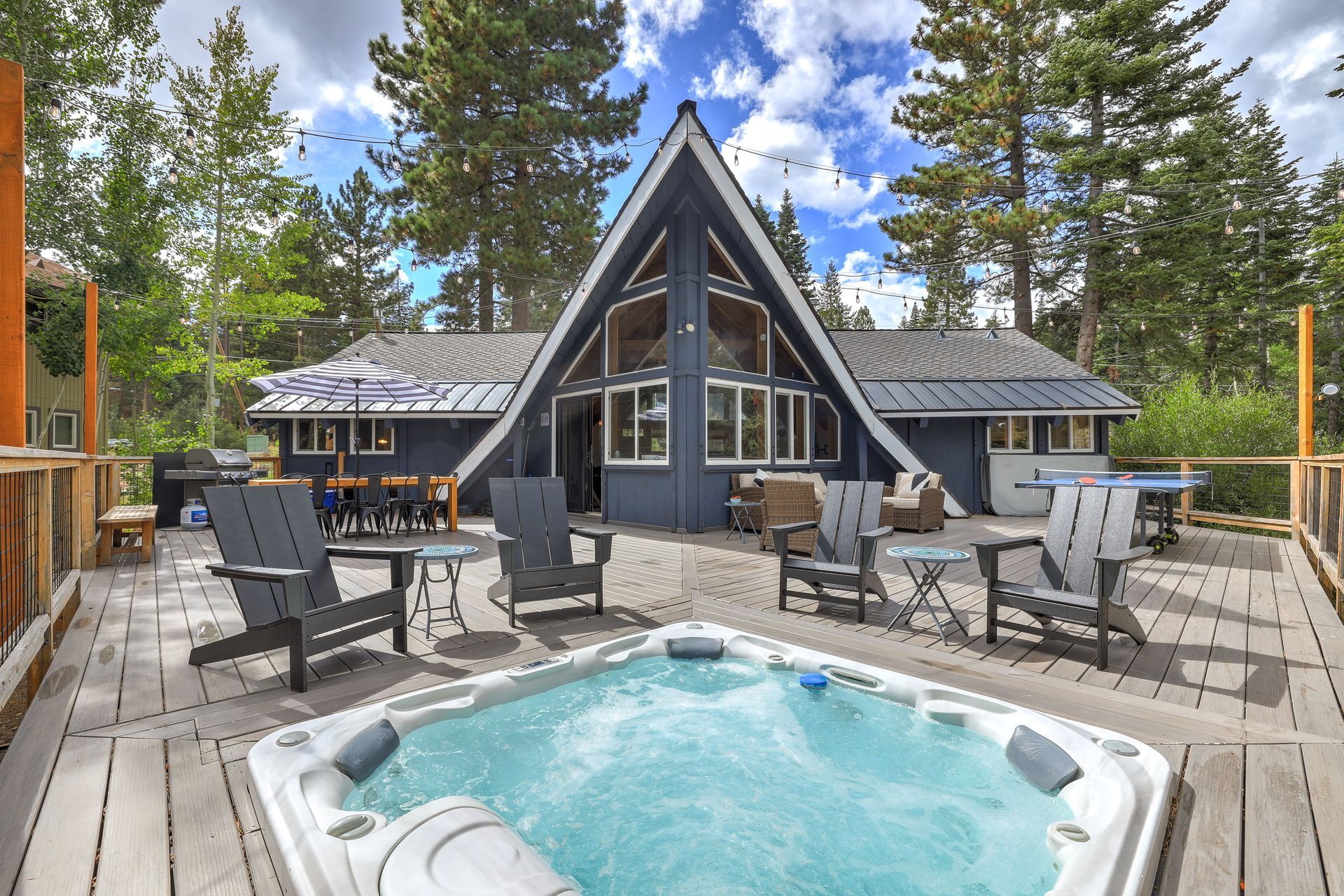A-frame cabin deck with hot tub, outdoor seating, and pine trees under a blue sky
