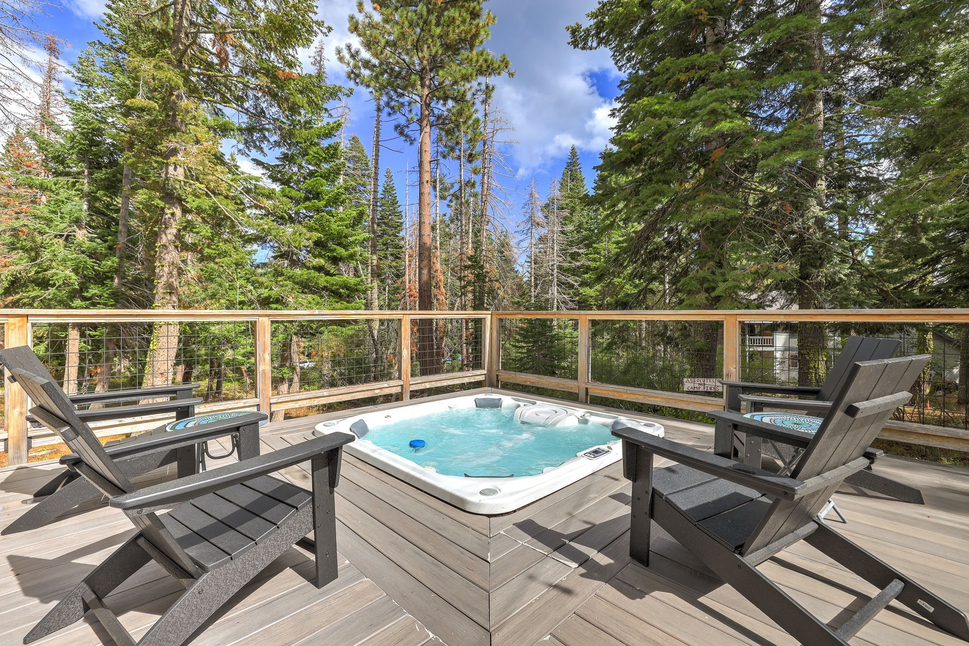 Hot tub on a wooden deck with Adirondack chairs and forest trees beyond a railing