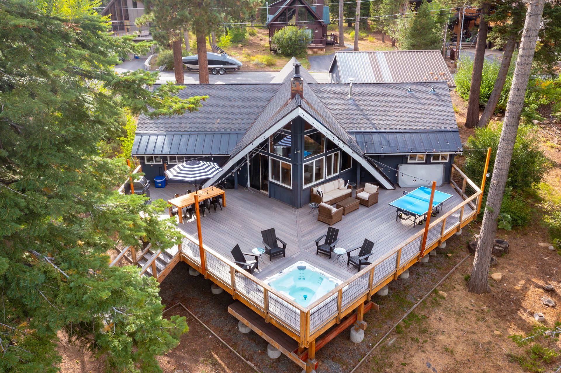 Aerial view of a rustic cabin deck with hot tub, patio furniture, and pine trees surrounding it.