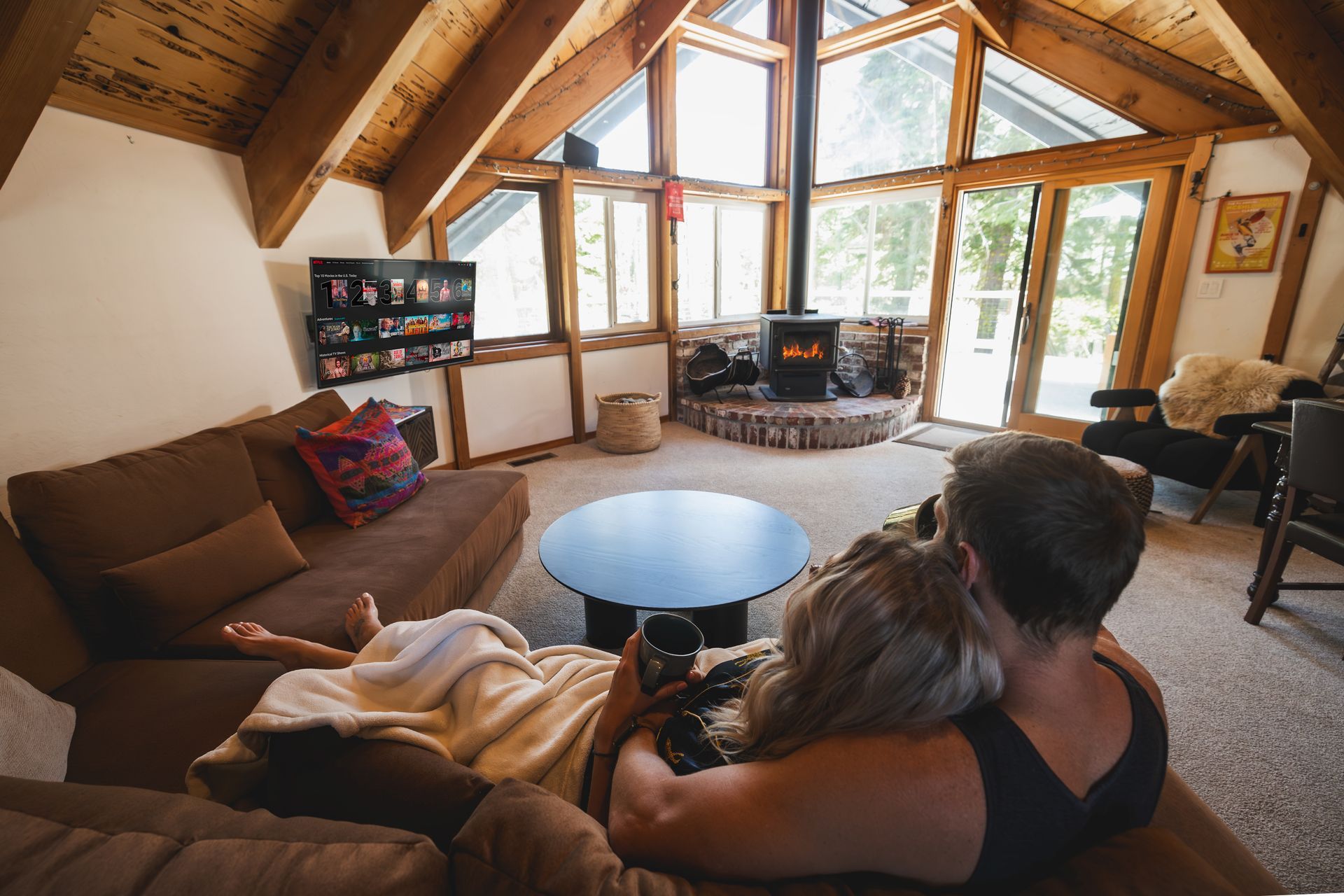 Cozy cabin living room with a couple on a couch facing a large fireplace and panoramic windows