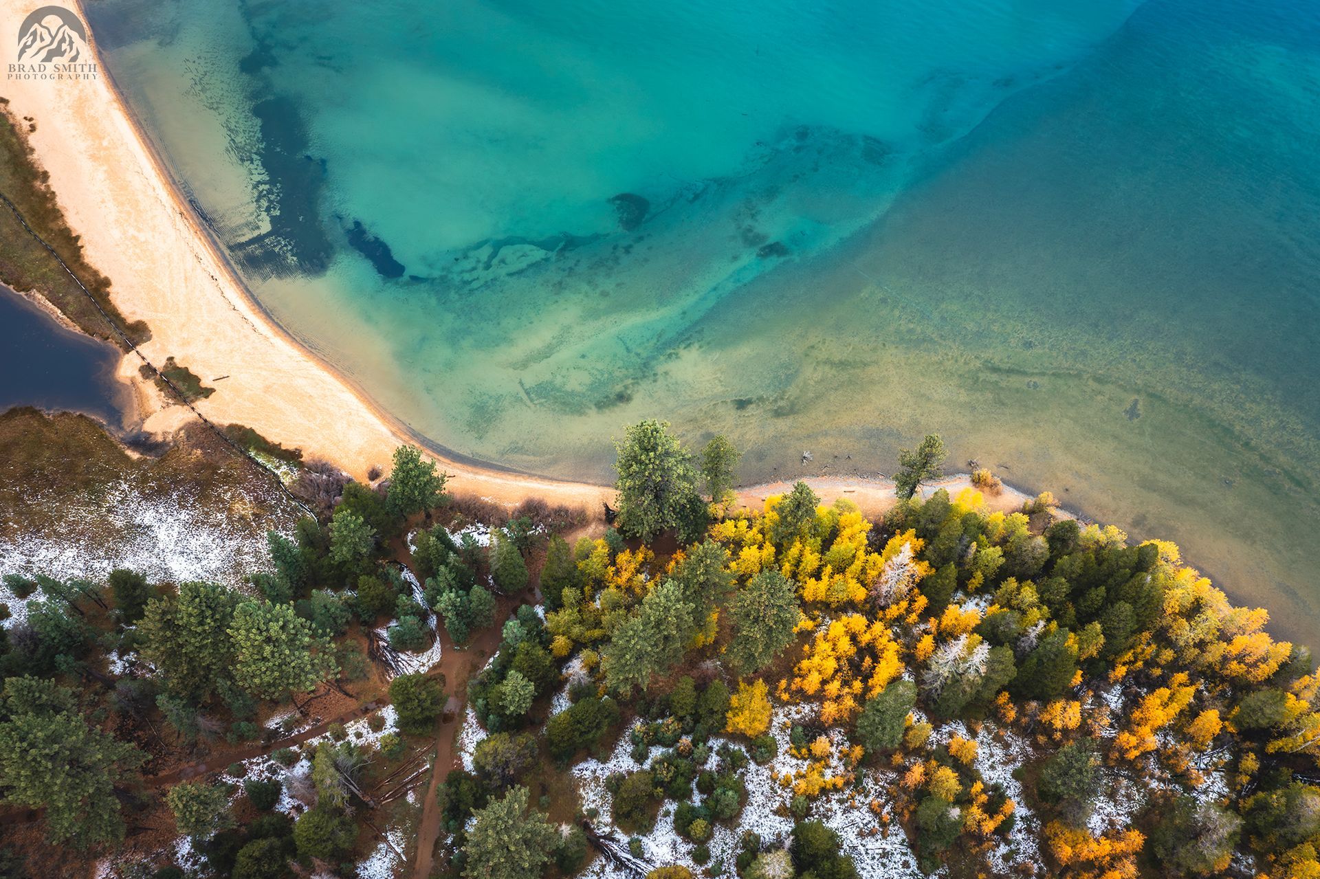 Aerial view of a sandy beach beside turquoise water and a forest with autumn-colored trees