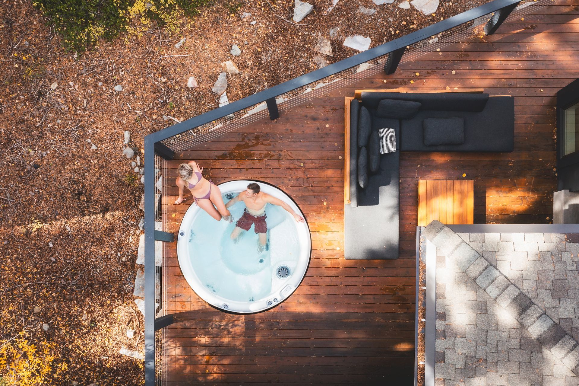 Person relaxing in a white hot tub on a wooden deck beside a black outdoor sofa.
