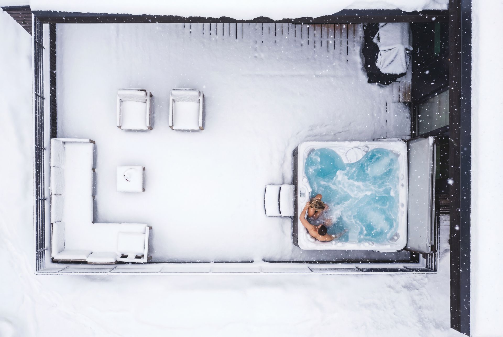 Snow-covered rooftop hot tub with one person soaking in steaming blue water