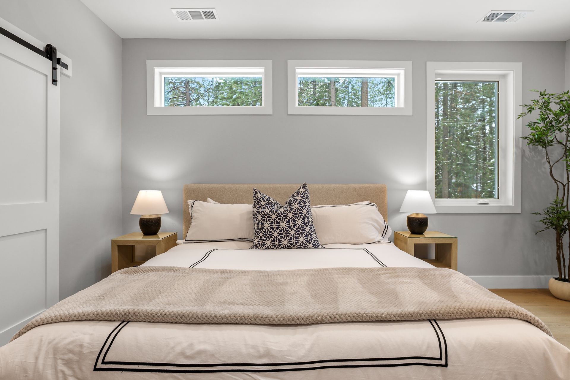 Modern bedroom with a beige bed, nightstands, lamps, and three windows on a light gray wall