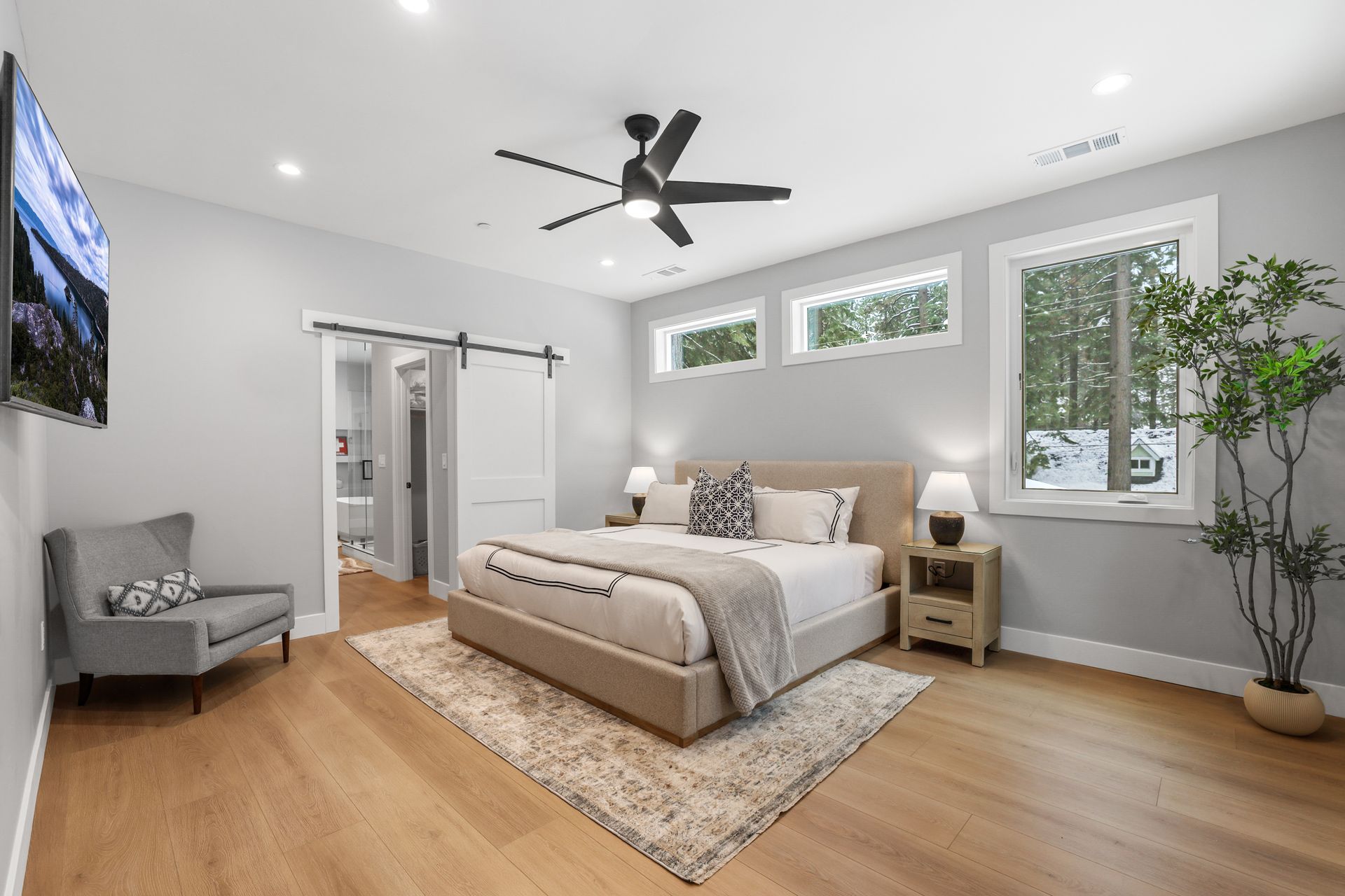 Bright modern bedroom with a gray bed, hardwood floors, ceiling fan, and large windows.