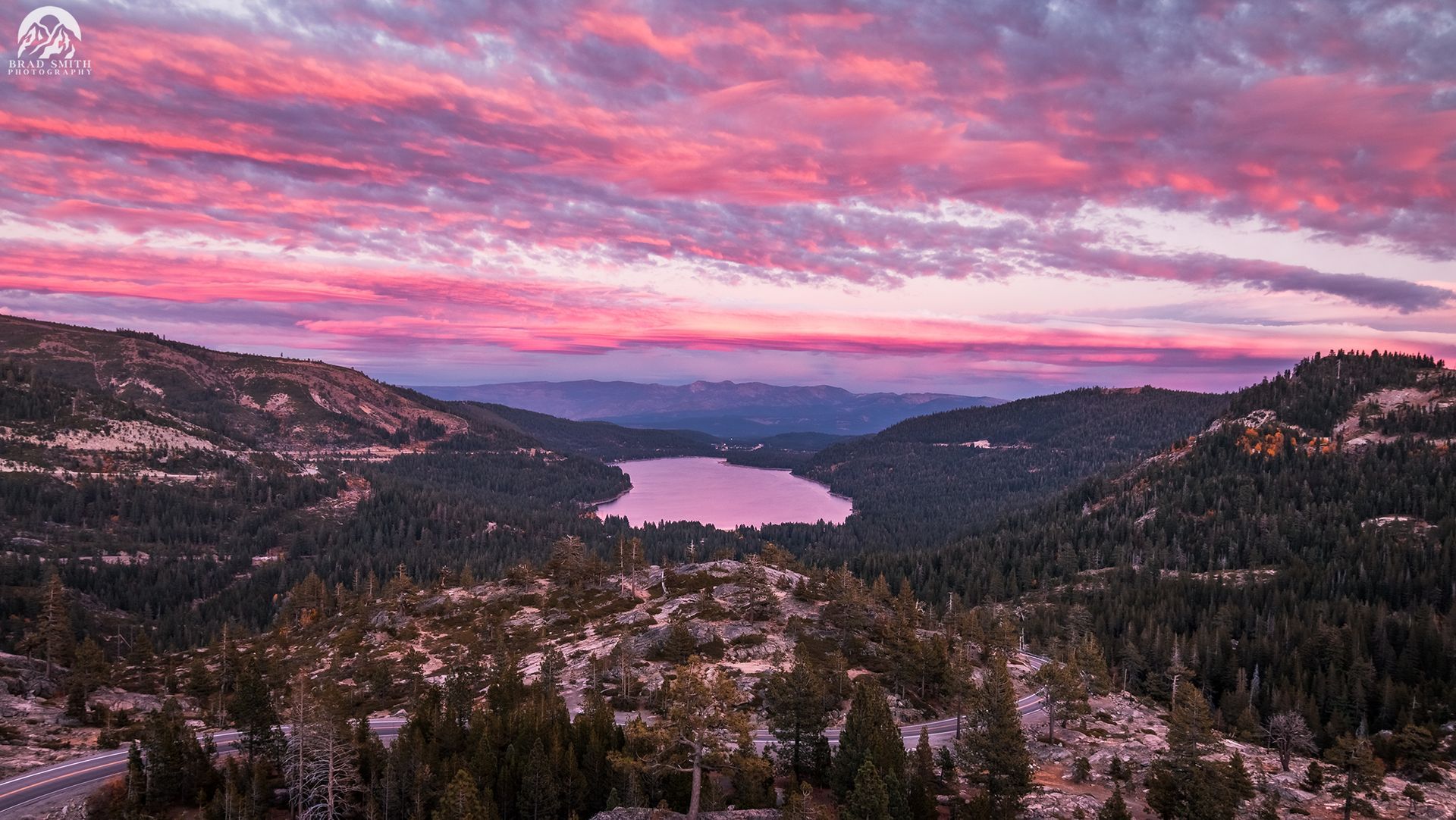 Mountain valley with a lake at sunset under pink clouds and forested hills.