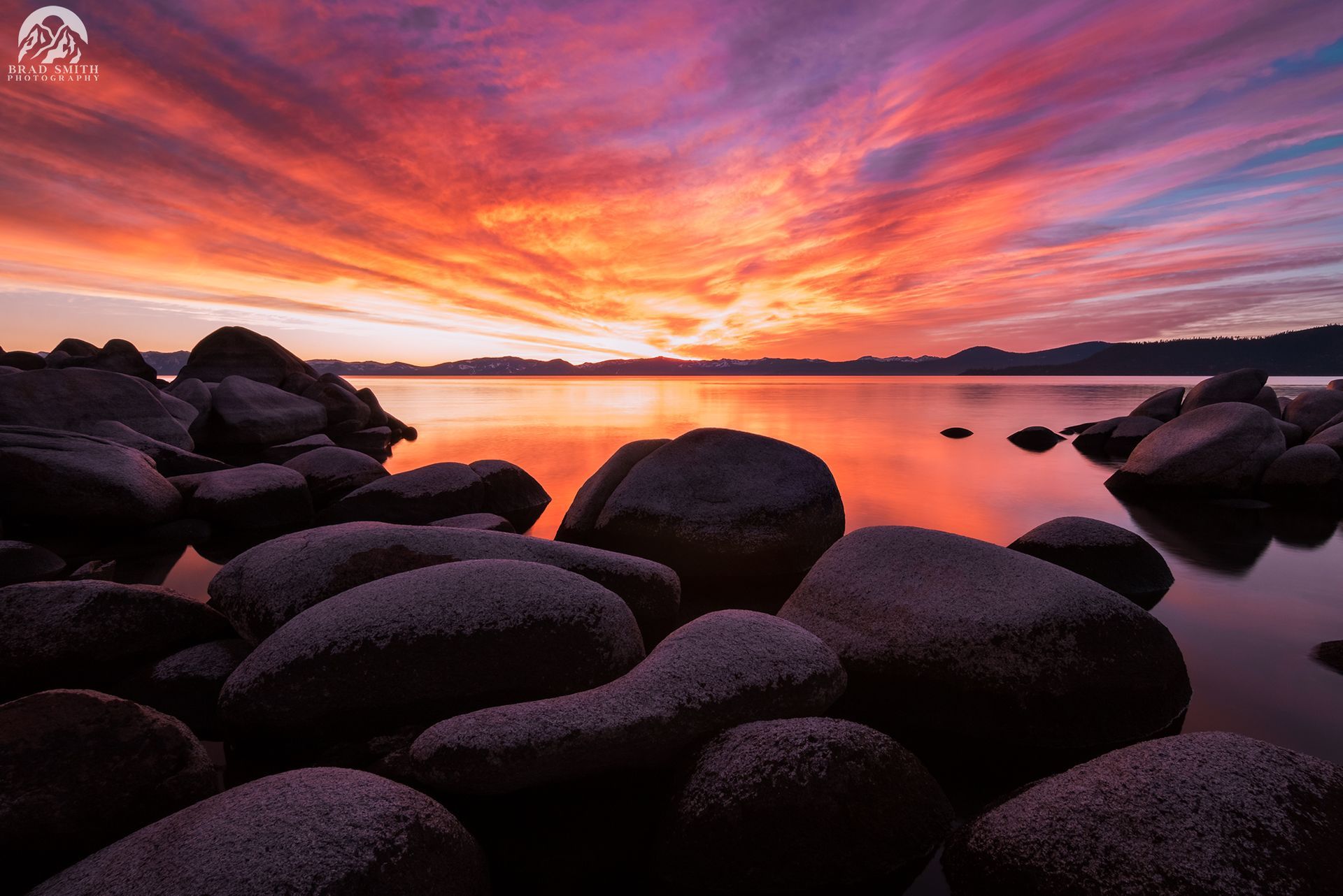 Sunset over calm water with dark rocky shoreline and vivid orange-purple clouds