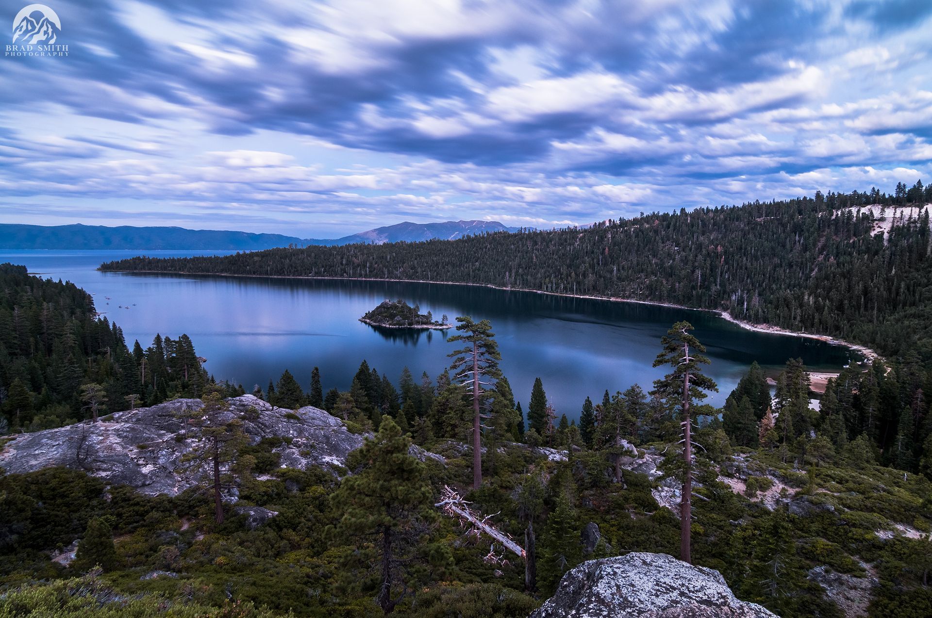 Lake surrounded by pine forest and rocky shoreline under a cloudy blue sky