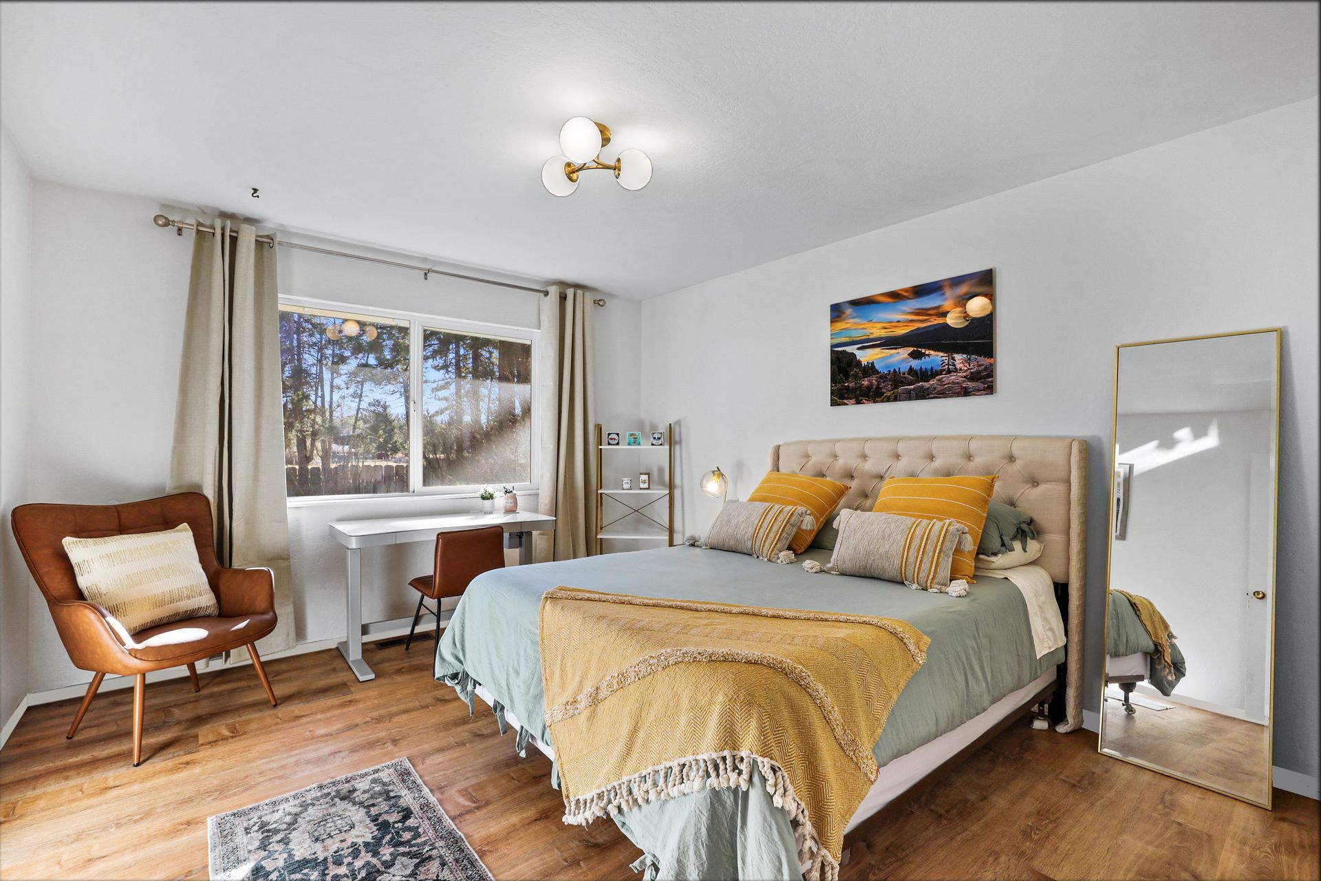Bright bedroom with a bed, chair, mirror, window, and warm wood floors