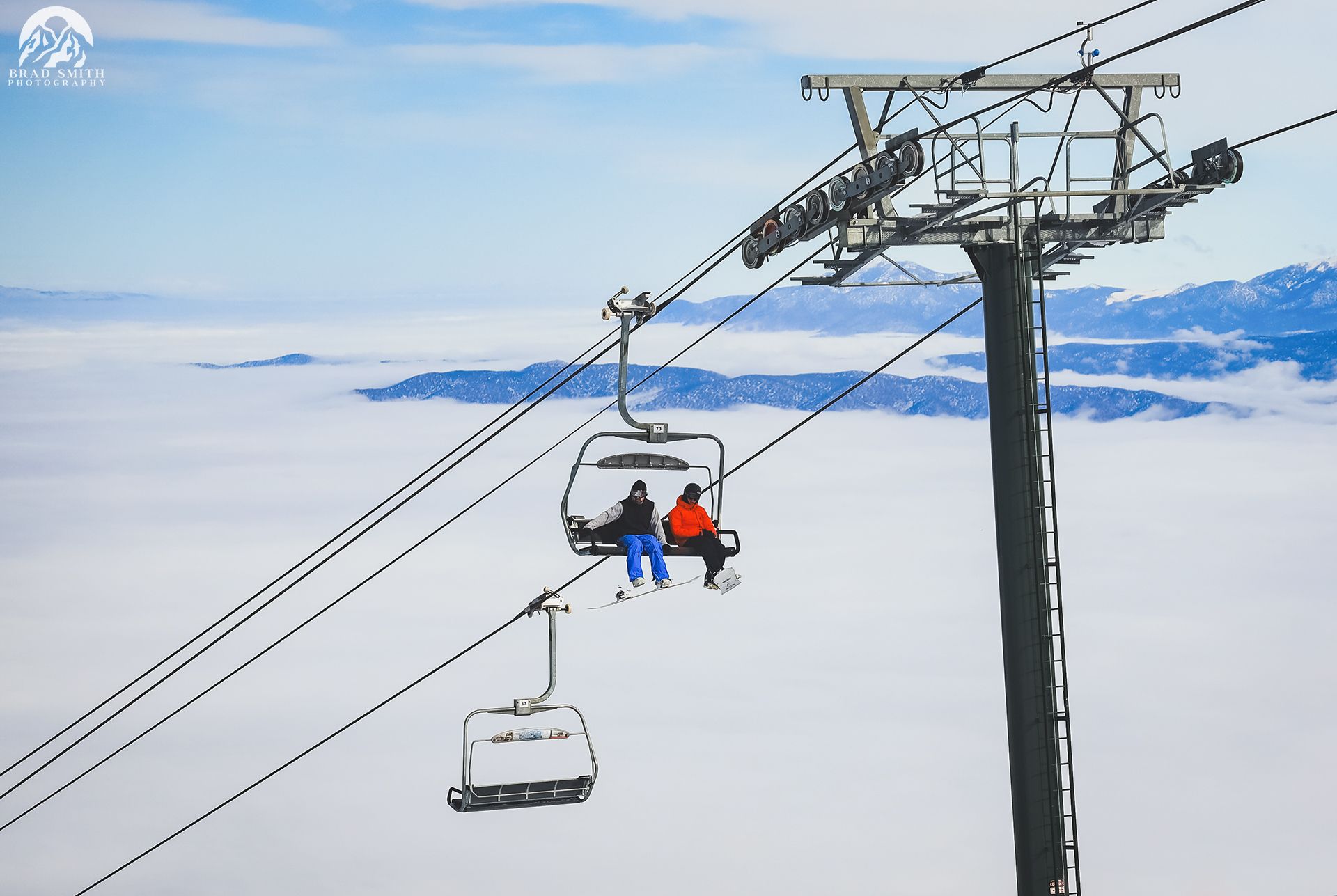 Two skiers on a chairlift above a snowy mountain cloudscape