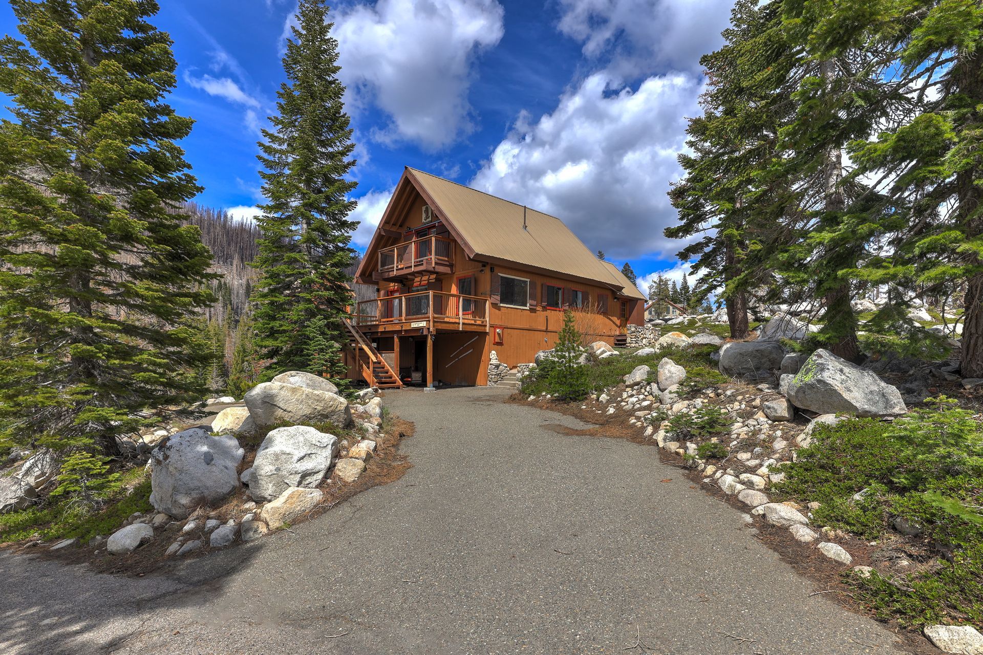 Wooden mountain lodge along a gravel path, framed by pine trees and rocks under a blue sky
