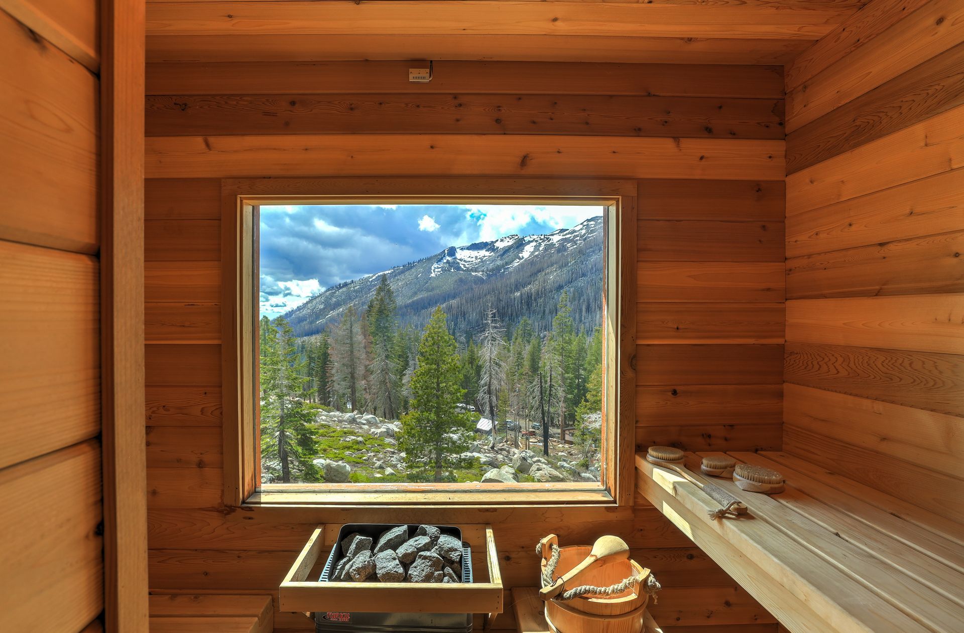 Wooden sauna interior with bench and stones, framing a snowy mountain and forest view through the window