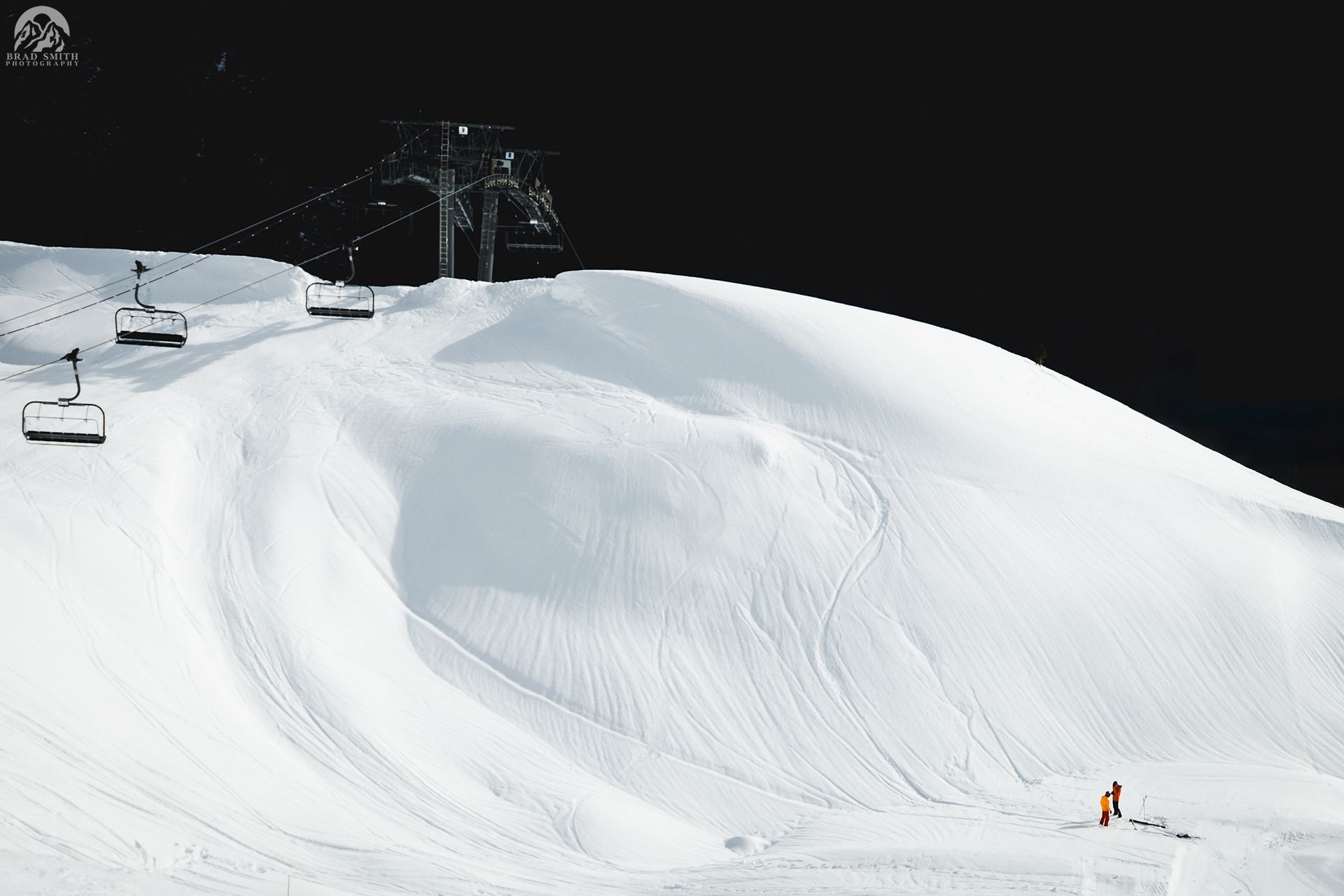 Snow-covered ski slope with a lone skier on a bright white hill under a dark sky