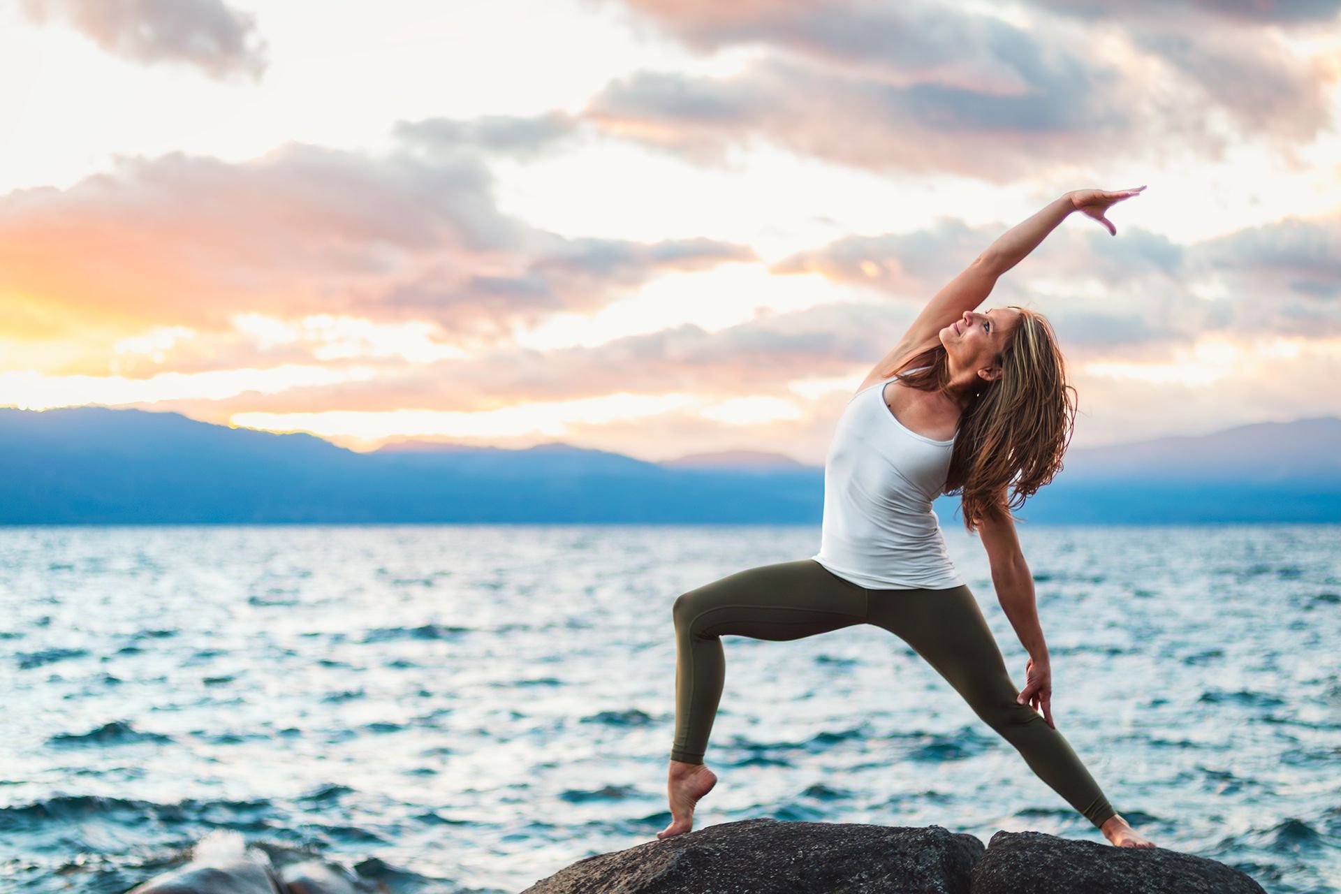 Person doing a yoga pose on a rocky shore at sunset, with ocean and colorful clouds behind.