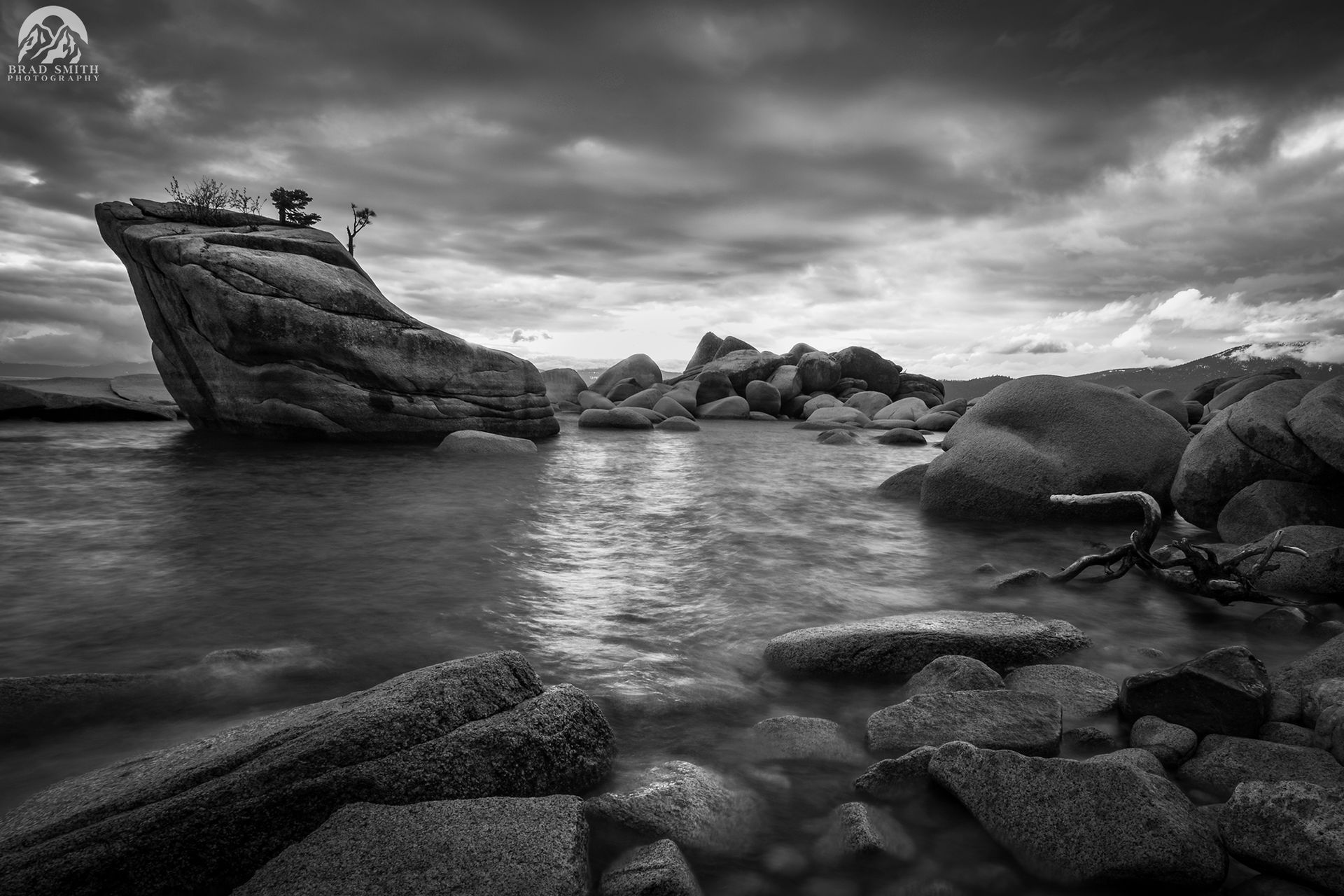 Black-and-white seascape with a rocky shore, calm water, and a clouded sky at sunset