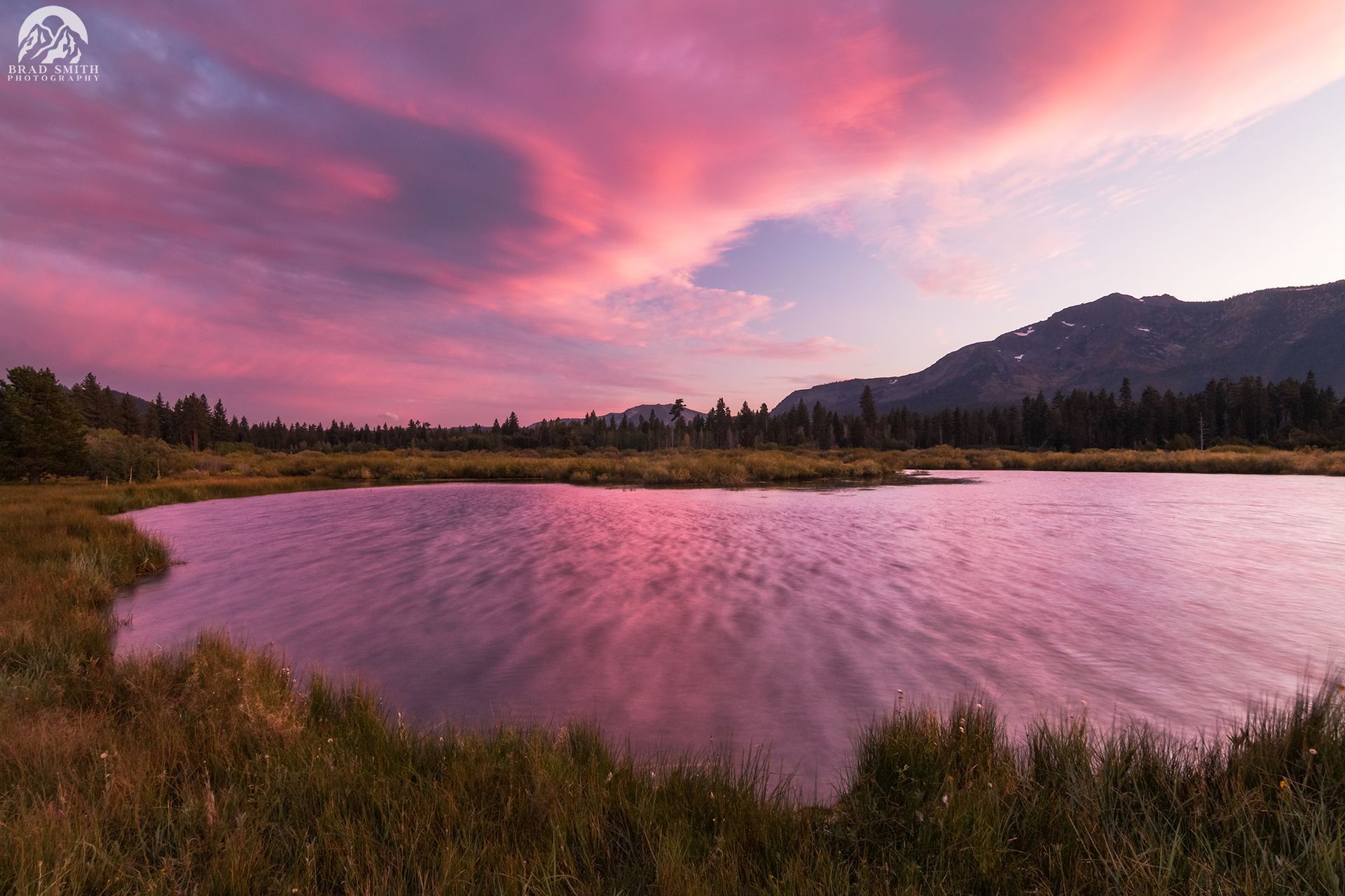 Pink sunset clouds reflected in a calm lake, with mountains and trees along the shore