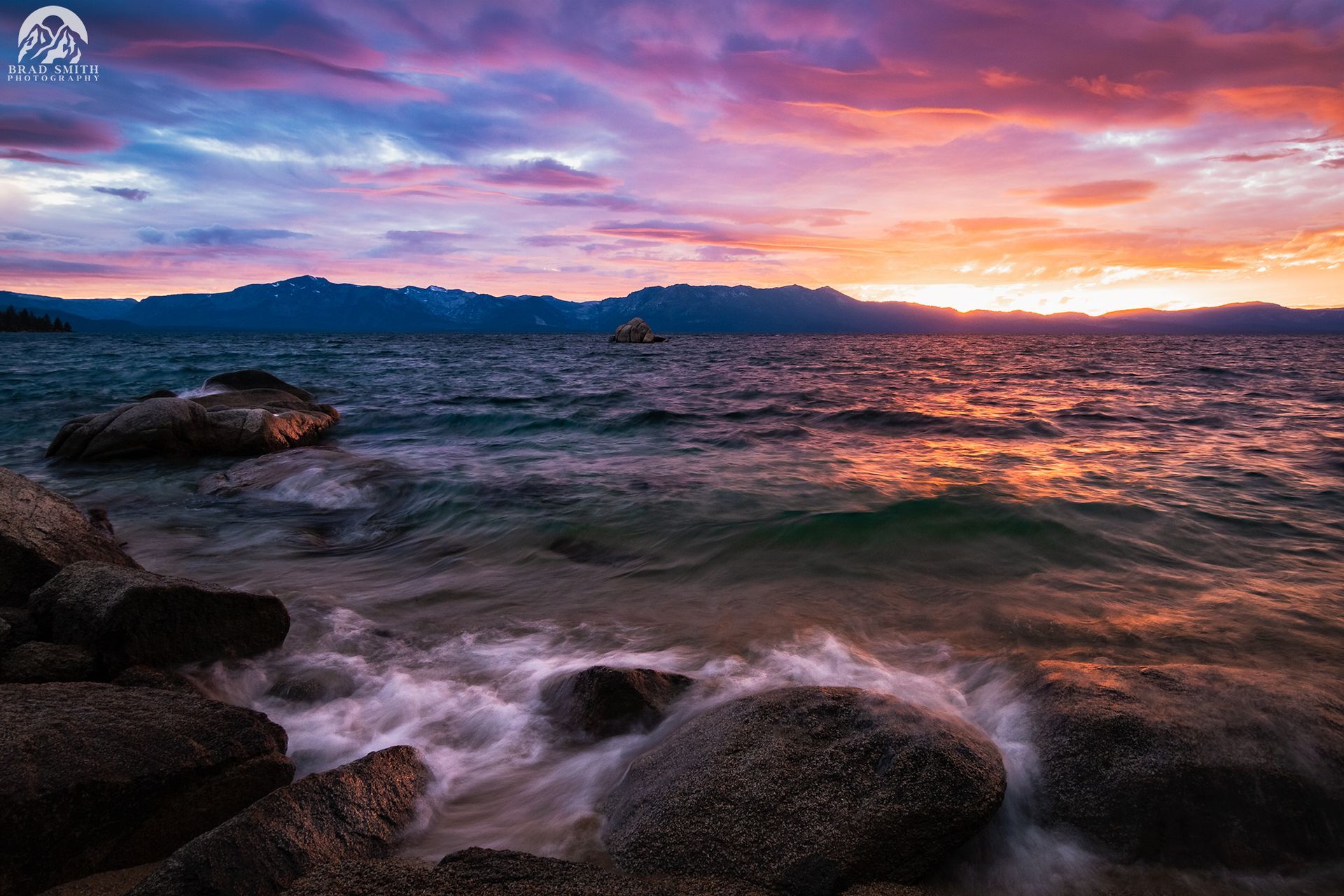 Rocky shoreline at sunset with waves and colorful clouds over distant mountains