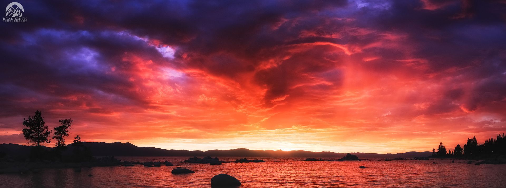 Vivid red-orange sunset over calm water with dark shoreline silhouettes and dramatic purple clouds