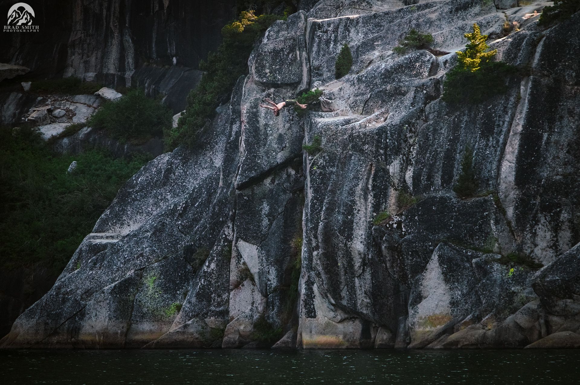Steep gray rock cliff with thin waterfalls and green moss above a dark pool