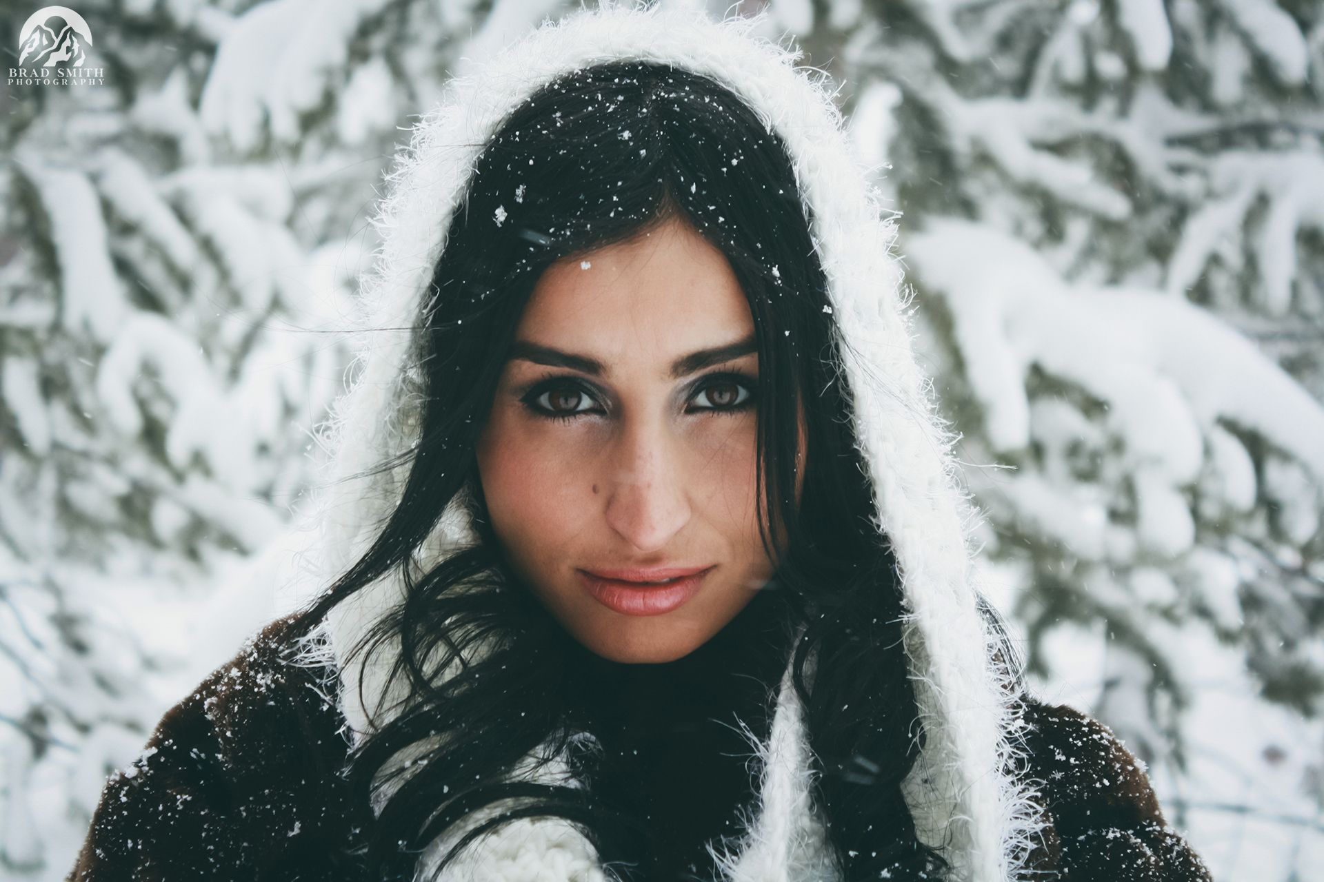 Woman with dark hair in a white hood, standing in snowy trees and looking at the camera.
