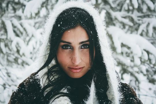 Woman in hooded winter coat smiles in snow, holding mittens up. Snowy forest in background.