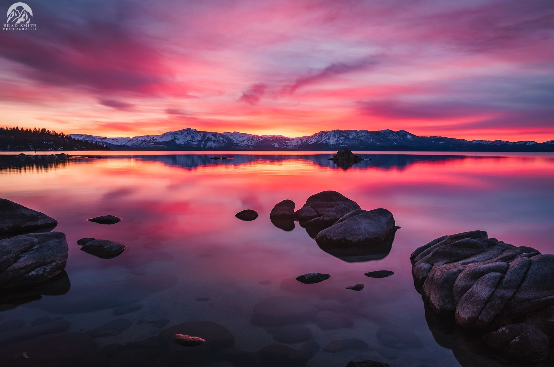 Pink sunset over a calm lake with rocky shoreline and distant snowy mountains