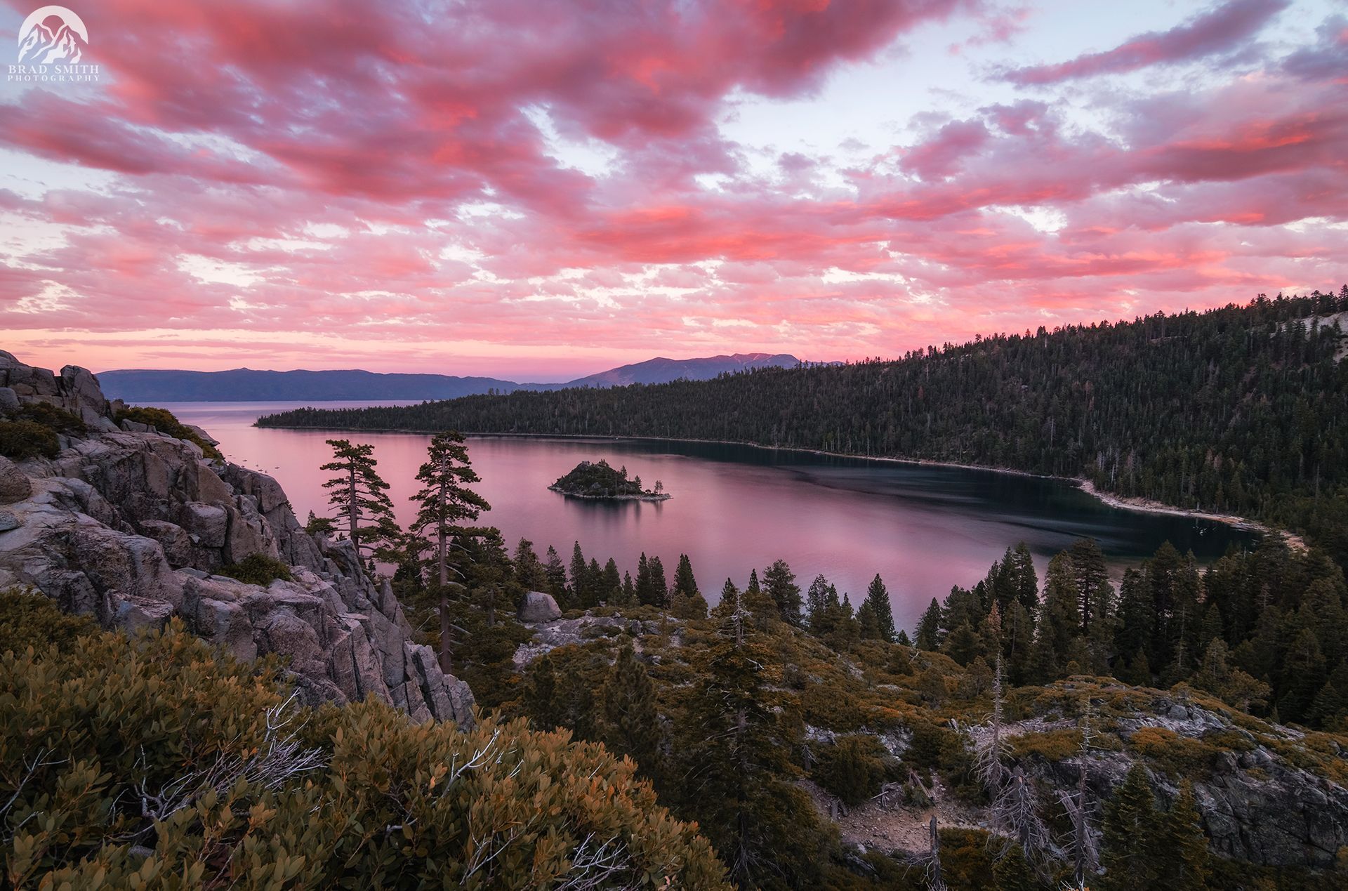 Pink sunset over a lake framed by pine-covered hills and rocky foreground