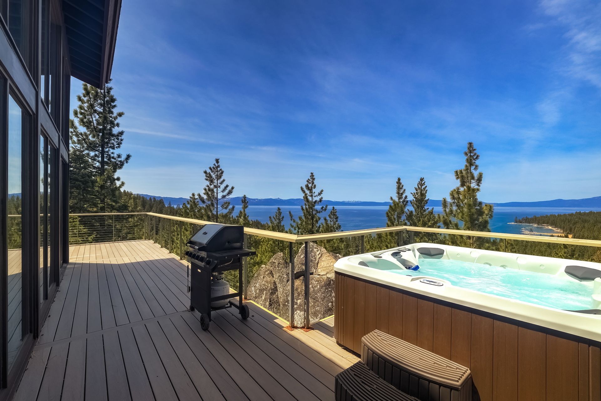 Wooden deck with hot tub and grill overlooking a blue lake and mountains under a clear sky