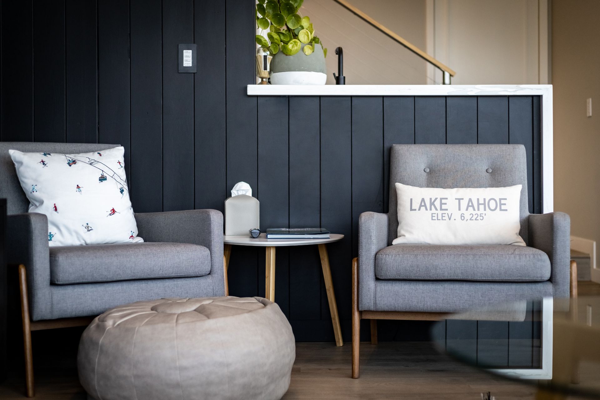 Cozy patio seating area with gray chairs, round ottoman, and a Lake Tahoe pillow against dark paneled walls