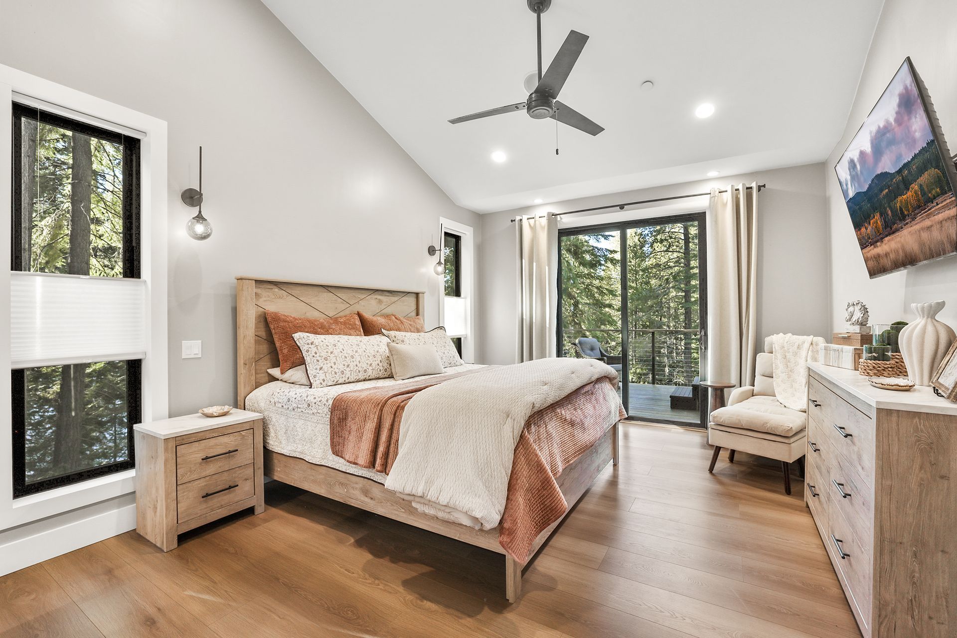Bright modern bedroom with a wooden bed, white walls, ceiling fan, and large windows overlooking trees