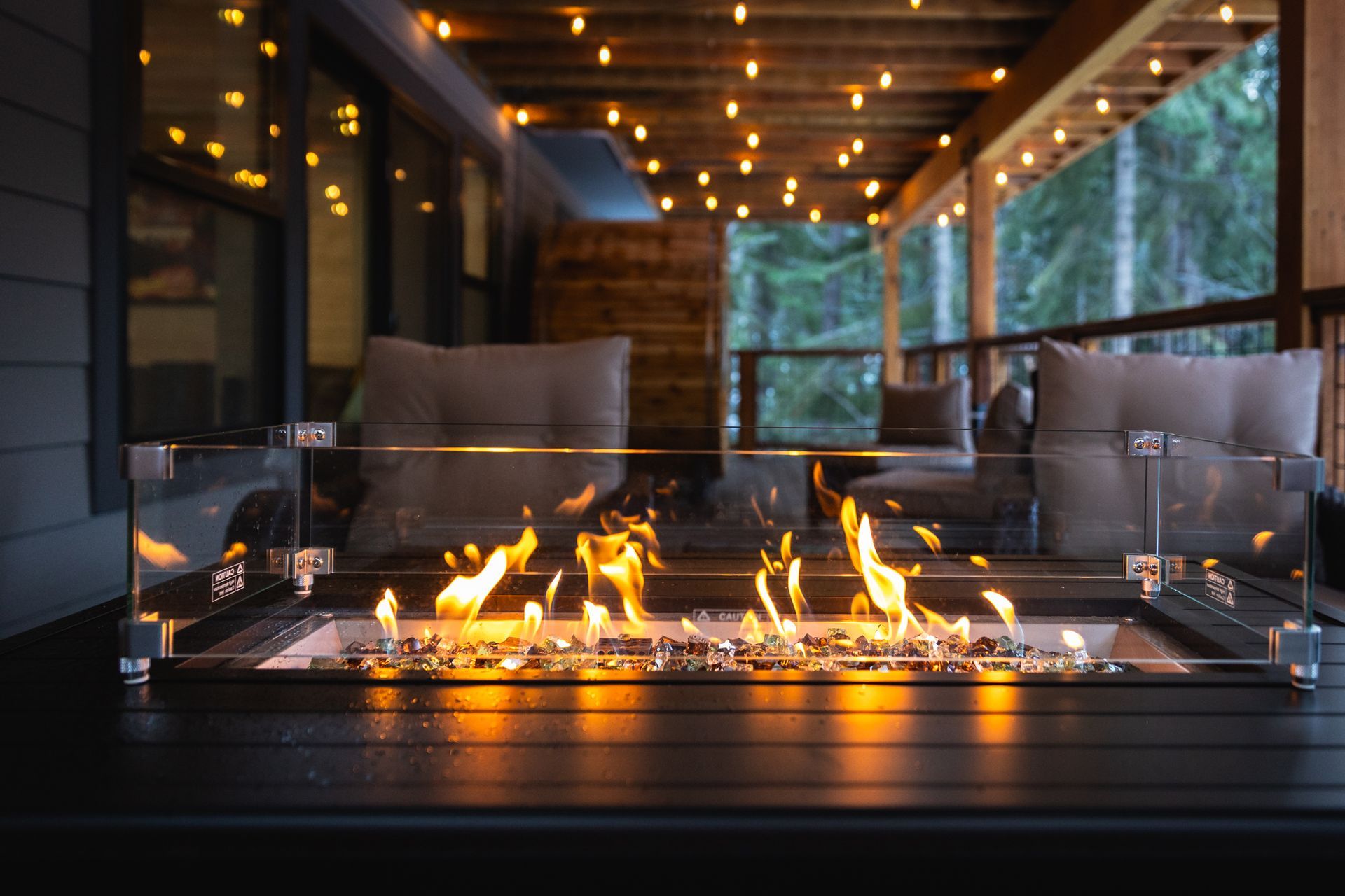 Modern patio with a glass fire pit, cushioned chairs, and warm string lights at dusk