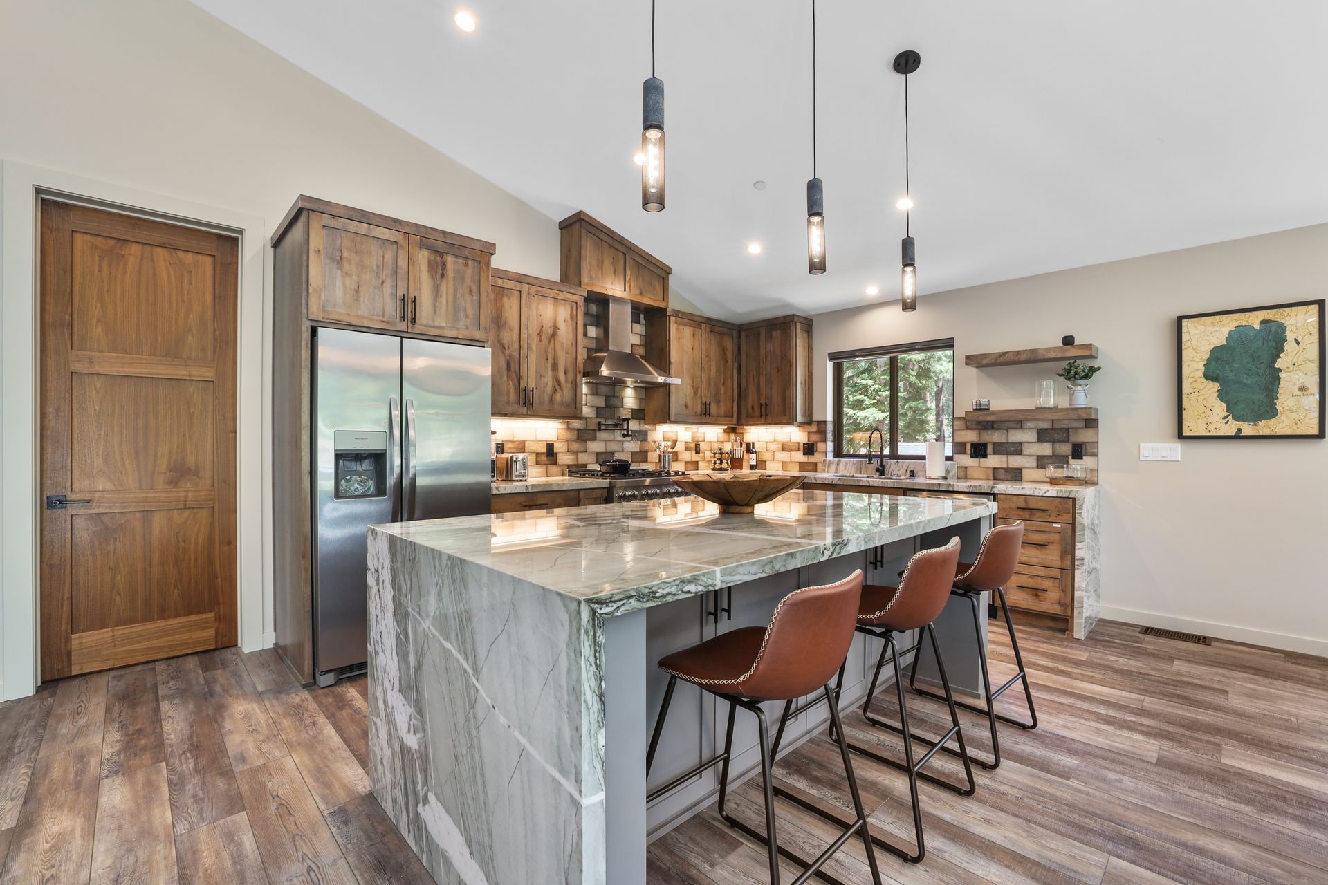 Modern kitchen with granite island, wood cabinets, stainless fridge, and four bar stools under pendant lights