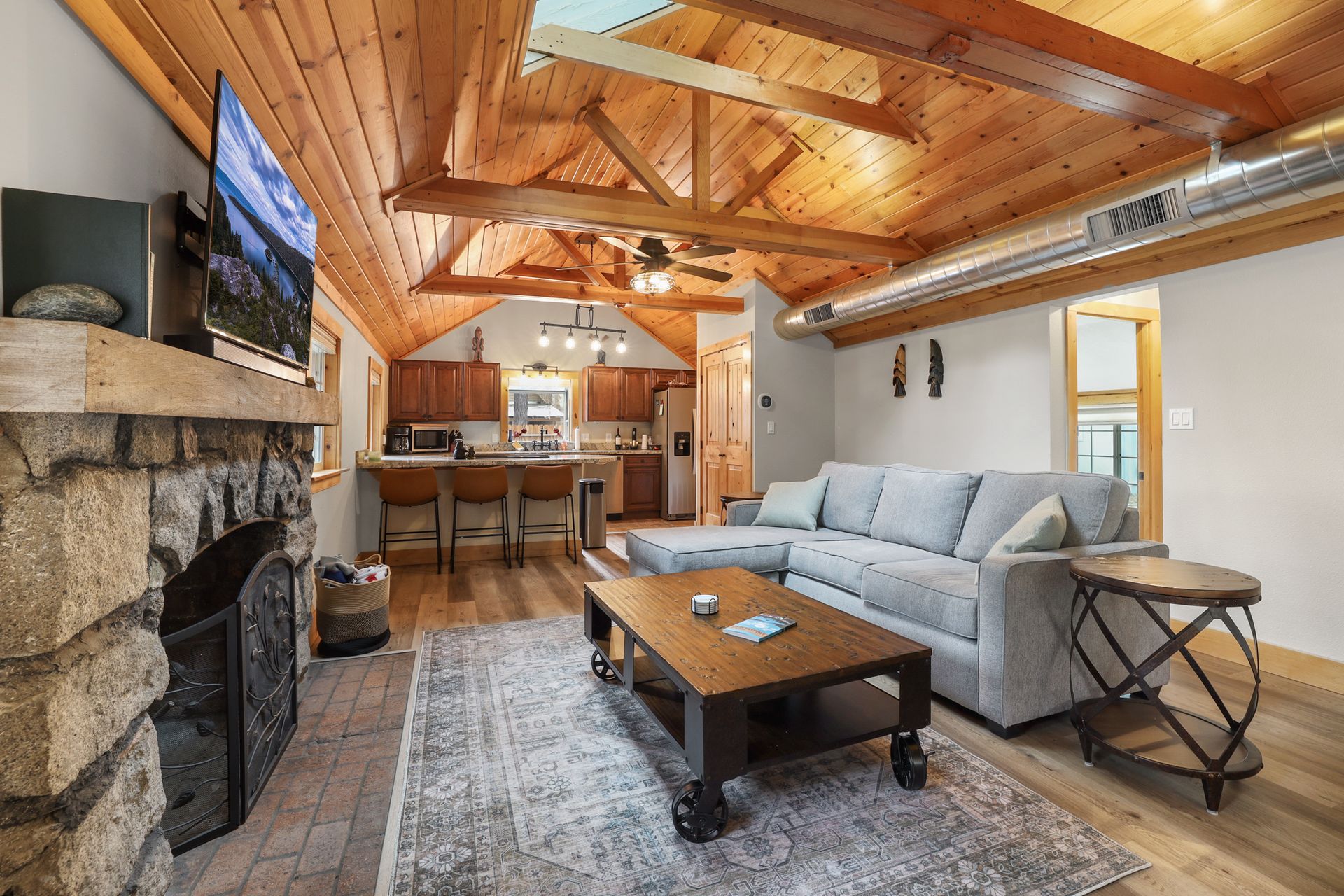 Cozy rustic living room with exposed wood beams, stone fireplace, gray sofa, and wooden coffee table